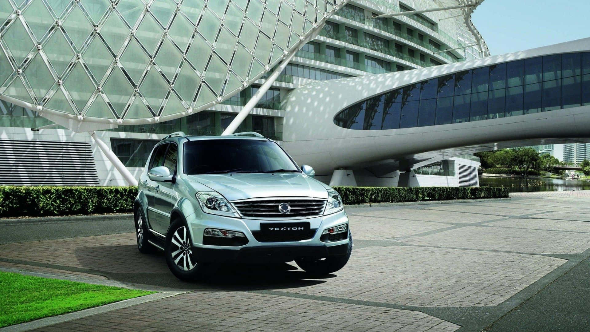 Ssangyong 桌布 壁紙 — 一輛銀色雙龍Tecton SUV停放在鋪砌的廣場上，前方是一座未來主義風格的玻璃和鋼結構建築，帶有弧形建築設計。