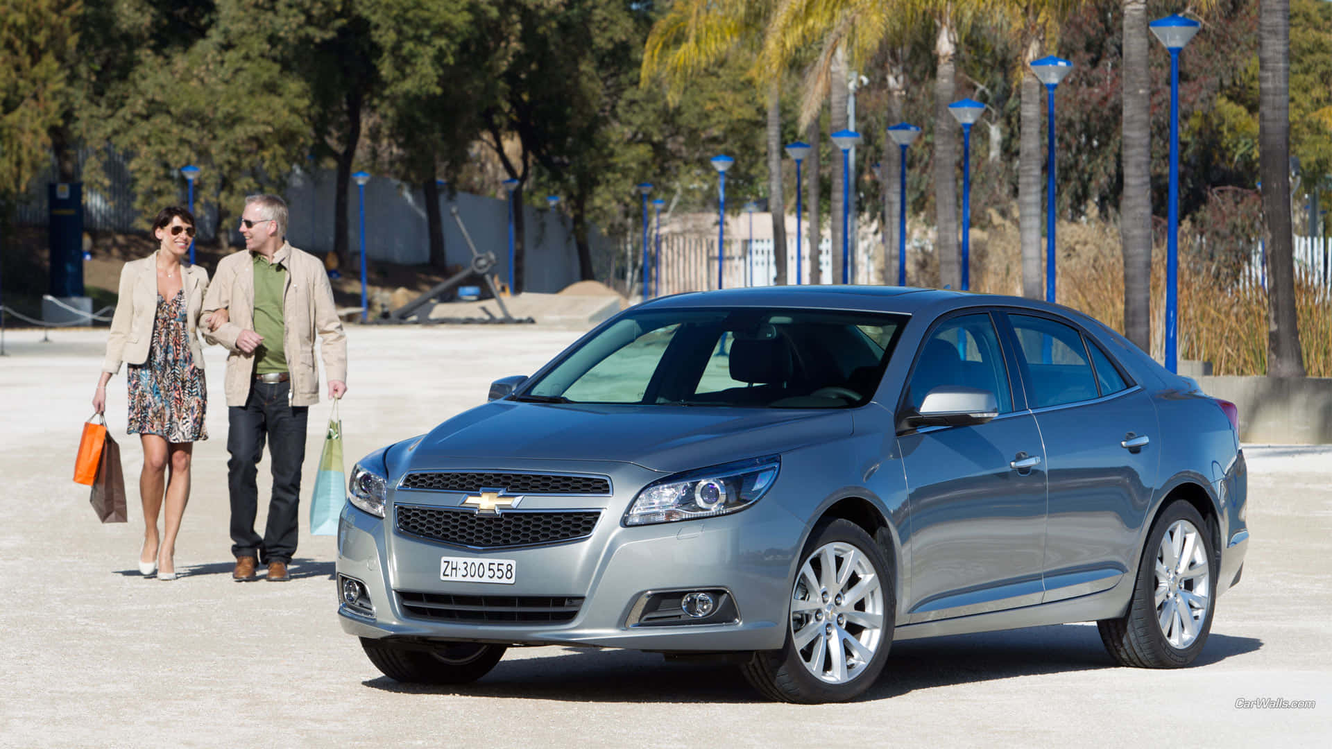 Chevy Malibu 桌布 壁紙 — 一輛銀色雪佛蘭轎車停放在陽光普照的廣場上，一對情侶提著購物袋走過。