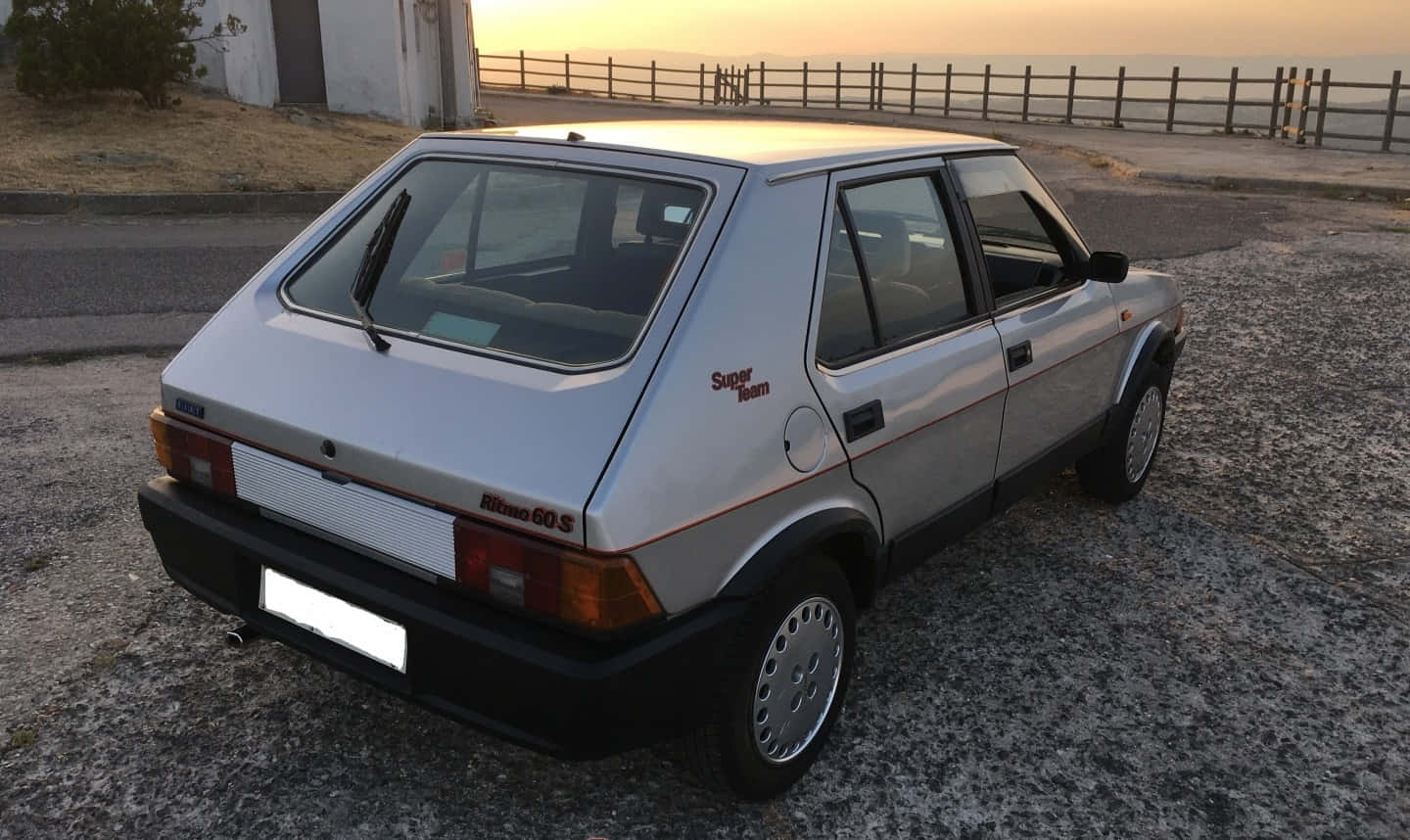 Fiat Ritmo 桌布 壁紙 — 一輛銀色飛雅特 Ritmo 60S 掀背車停在日落時分的碎石路上，背景有木柵欄和遠方的大海。