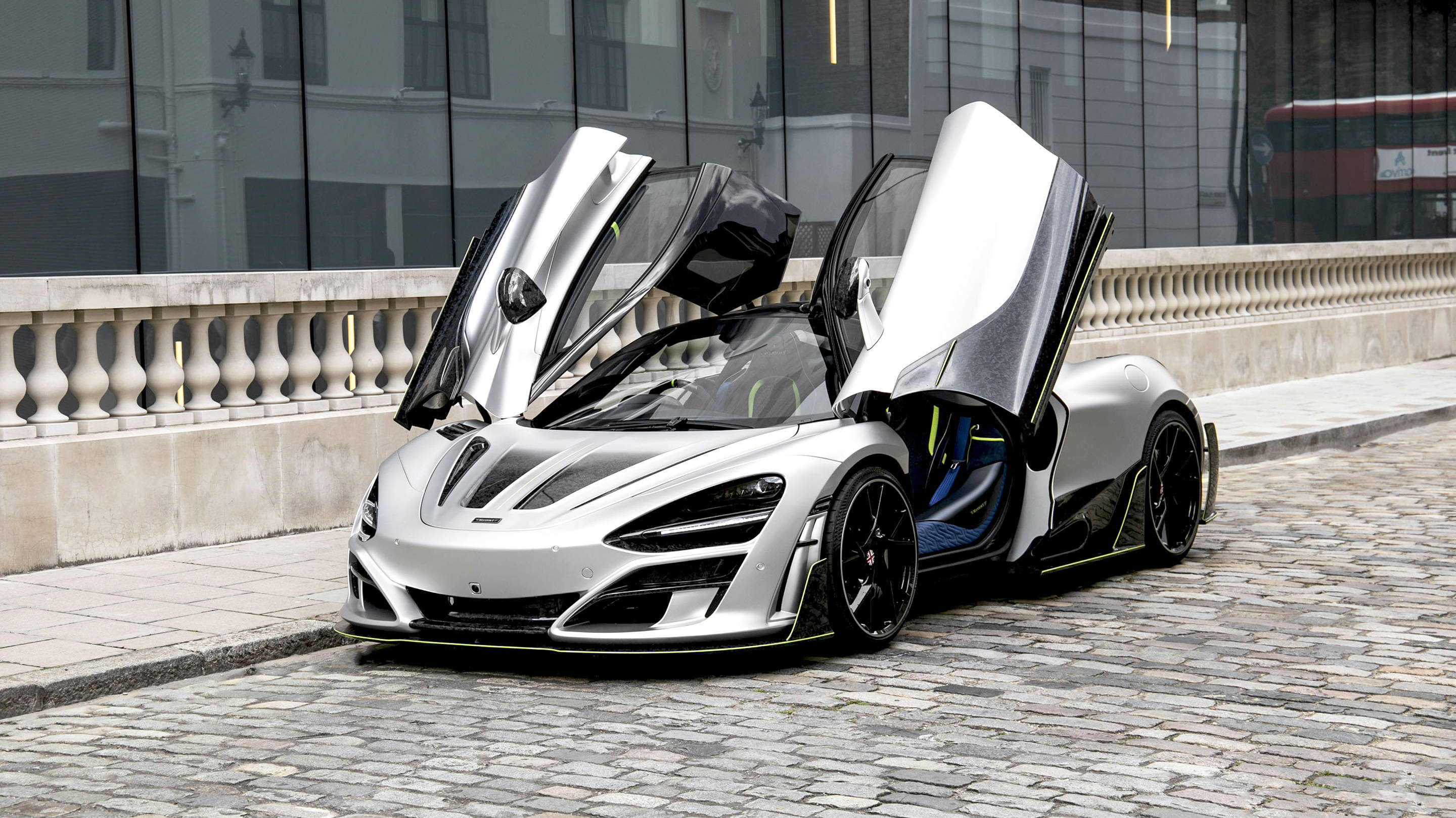麥拉倫 720S 桌布 壁紙 — 一輛銀色麥拉倫Senna，鷗翼車門打開，停在鵝卵石街道上，旁邊是現代玻璃建築。