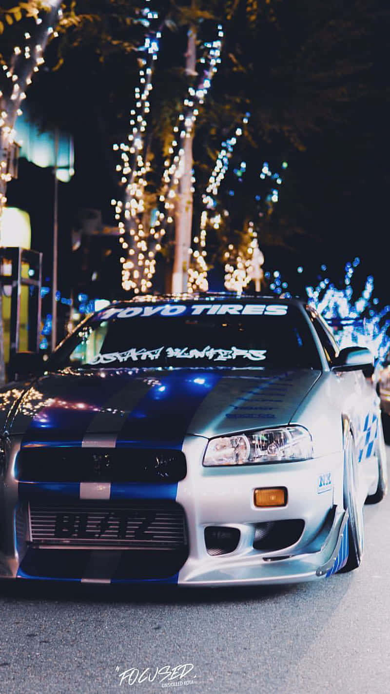 Paul Walker Skyline 桌布 壁紙 — 一輛銀藍色改裝跑車在夜間停泊在樹蔭街道上閃爍的燈串下。