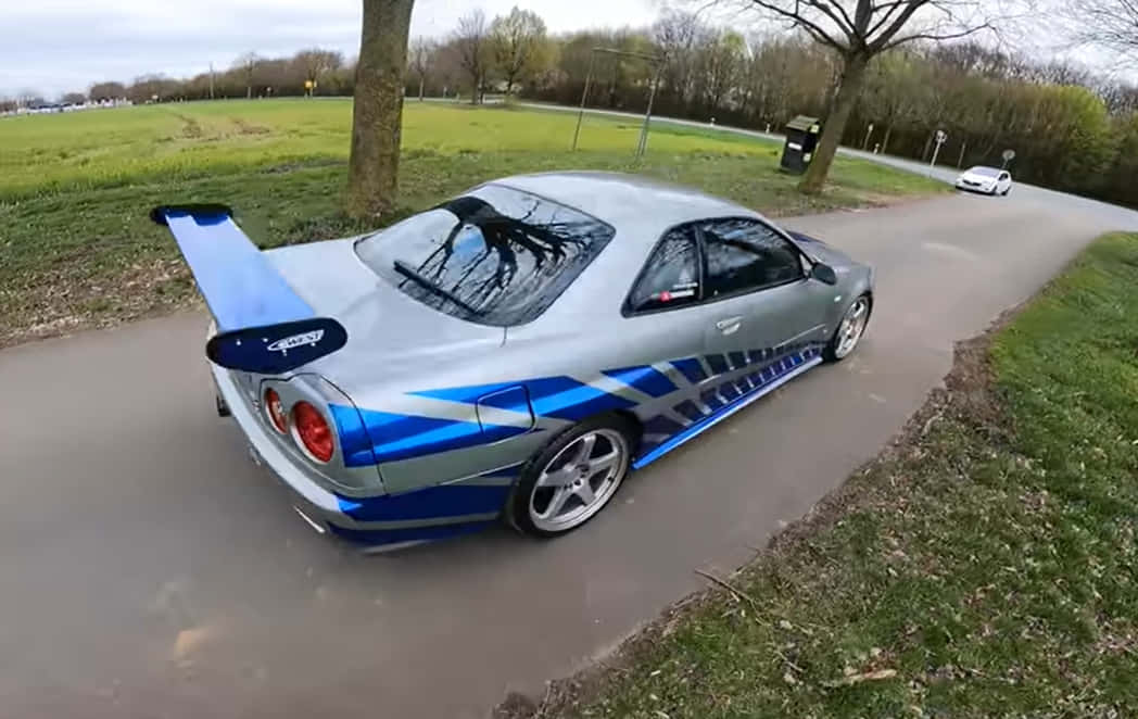 Paul Walker Skyline 桌布 壁紙 — 一輛銀藍色日產GT-R配備大型尾翼，在蜿蜒的公園道路上行駛，周圍環繞著綠草和光禿禿的樹木。