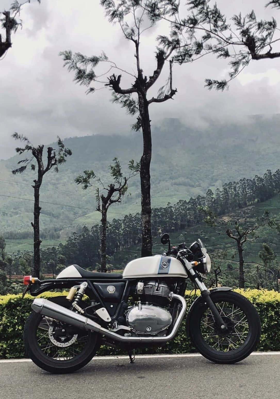 Royal Enfield Bullet 桌布 壁紙 — 一輛銀黑色經典機車停放在路邊，背景是霧氣瀰漫的翠綠山巒和稀疏樹木。