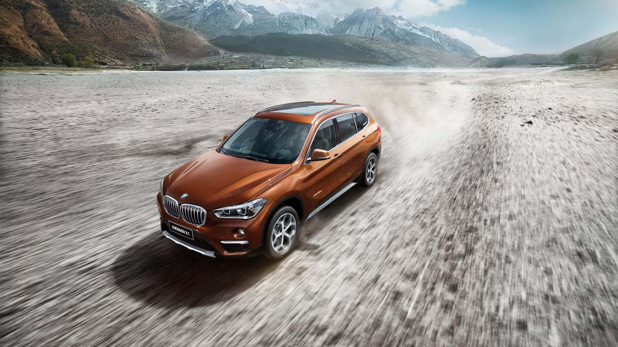 Bmw X1 桌布 壁紙 — 一輛銅色BMW X1 SUV在塵土飛揚的河床上疾馳，背景是白雪覆蓋的山脈。