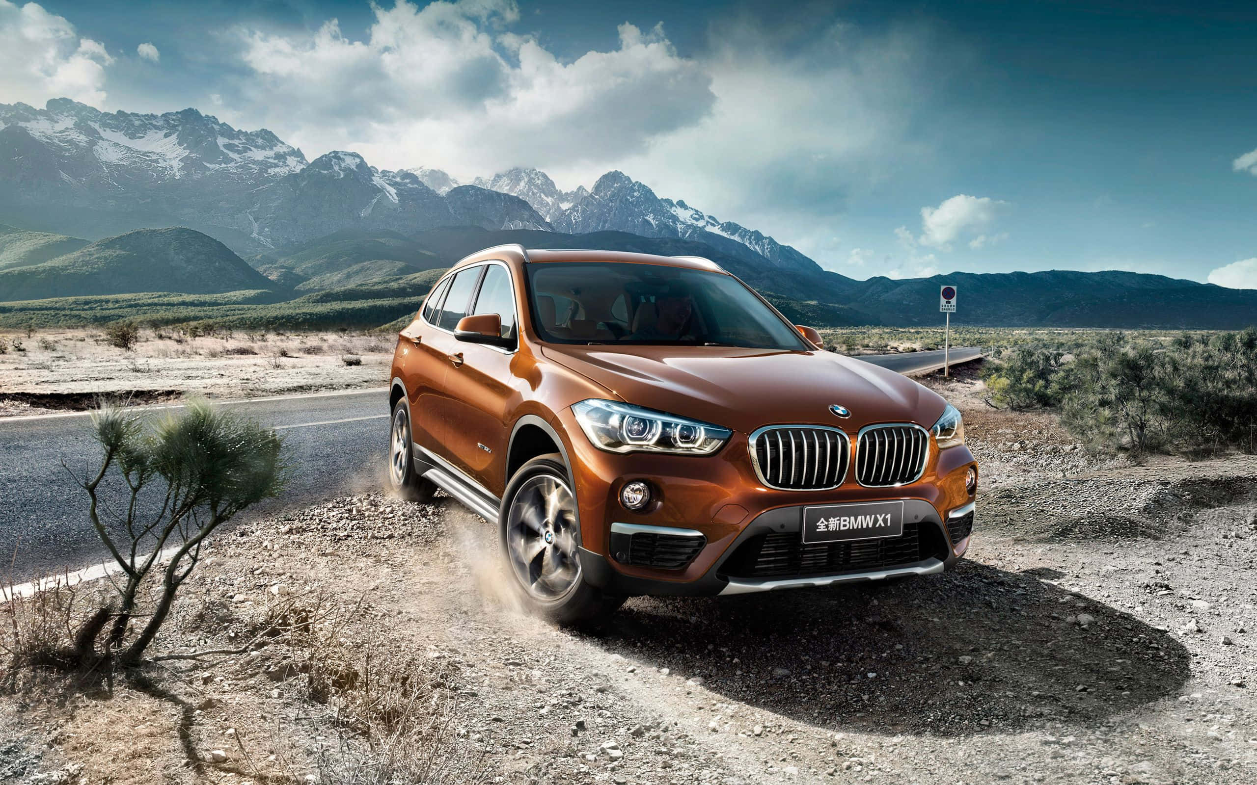 Bmw X1 桌布 壁紙 — 一輛銅色BMW X1 SUV在沙漠公路上行駛，背景是雲層密佈的天空下積雪覆蓋的山脈。