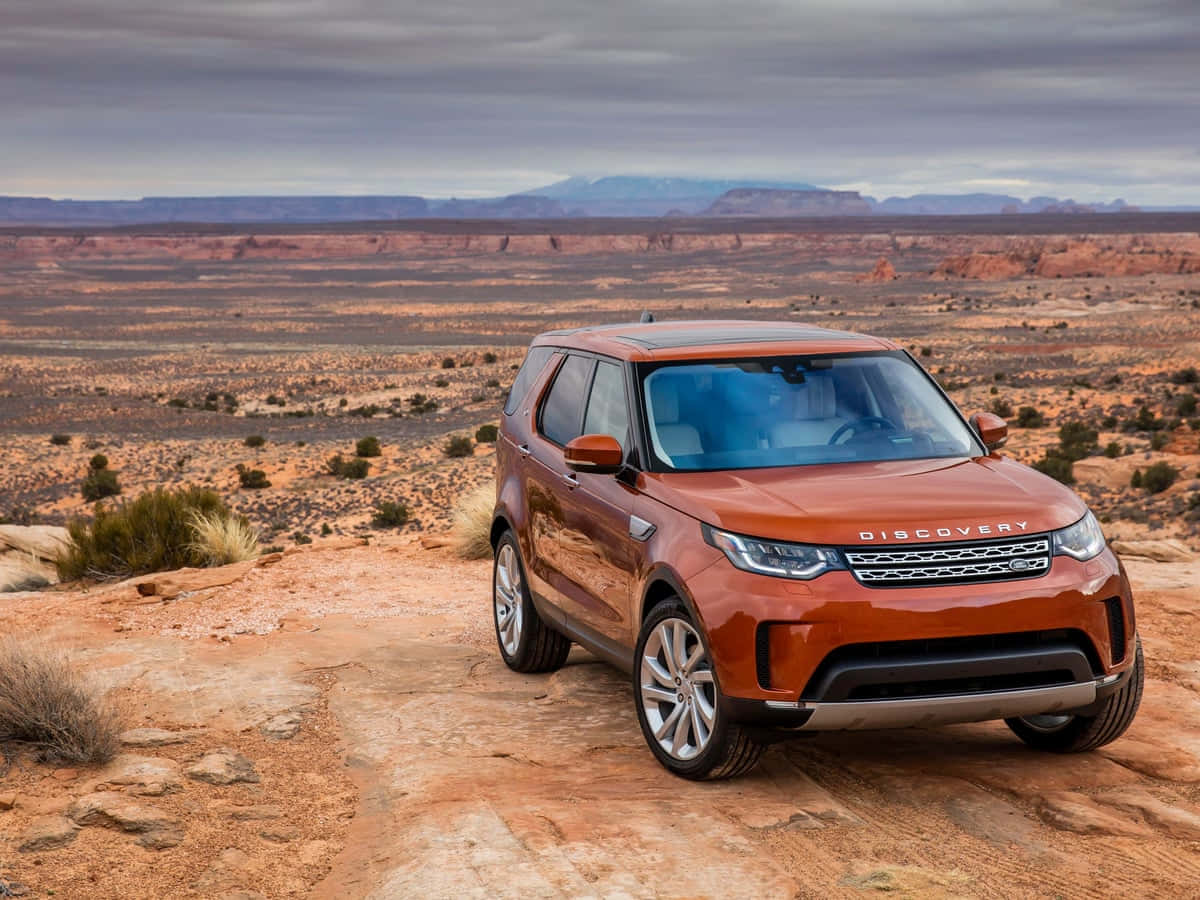 Land Rover Discovery 桌布 壁紙 — 一輛銅色的Land Rover Discovery停放在岩石沙漠小徑上，遠處有平頂山，天空多雲。