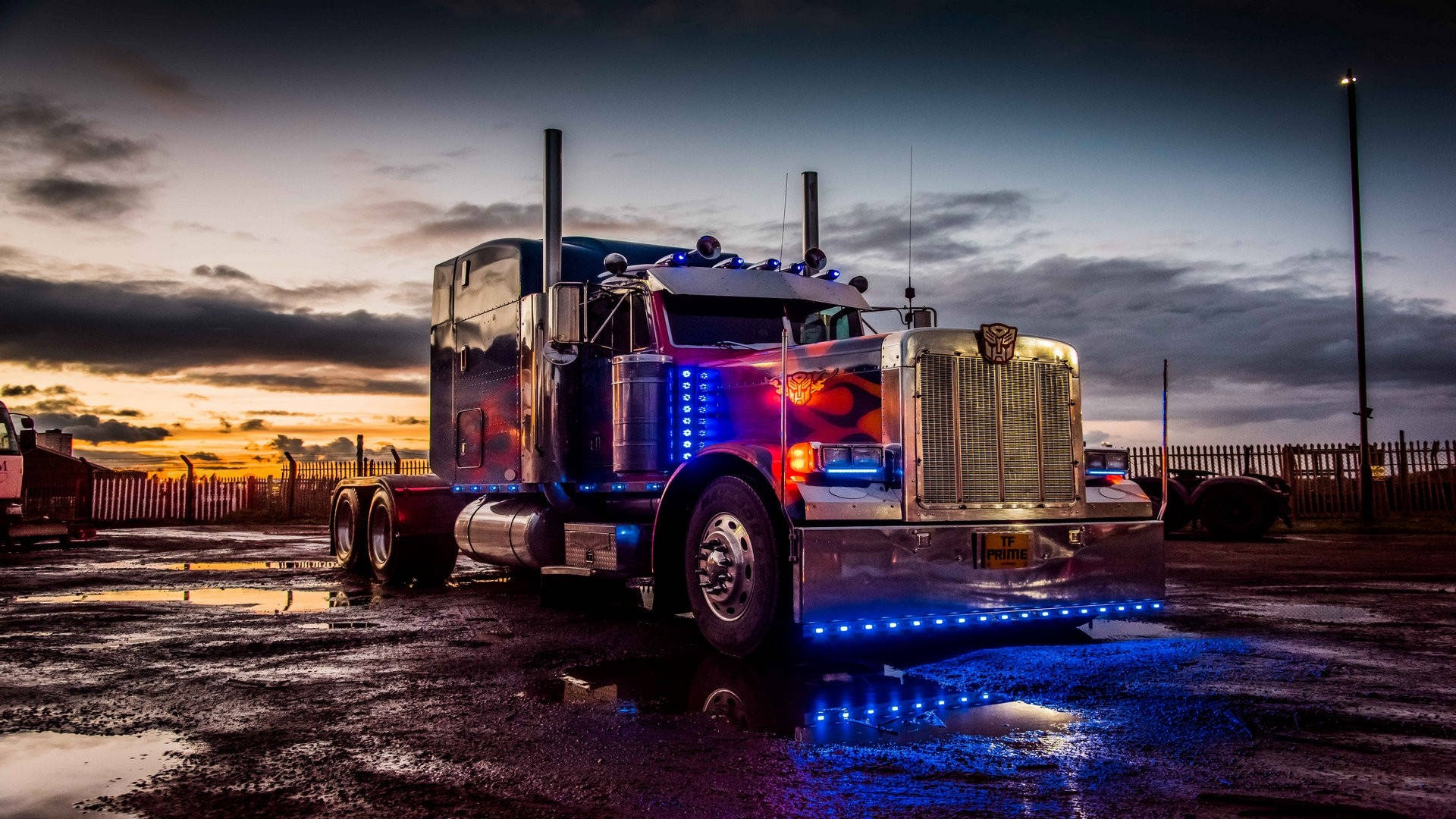 Trucks 桌布 壁紙 — 一輛鍍鉻半掛卡車在日落時分，多雲的天空下，藍色LED燈光映照在濕漉漉的路面上。