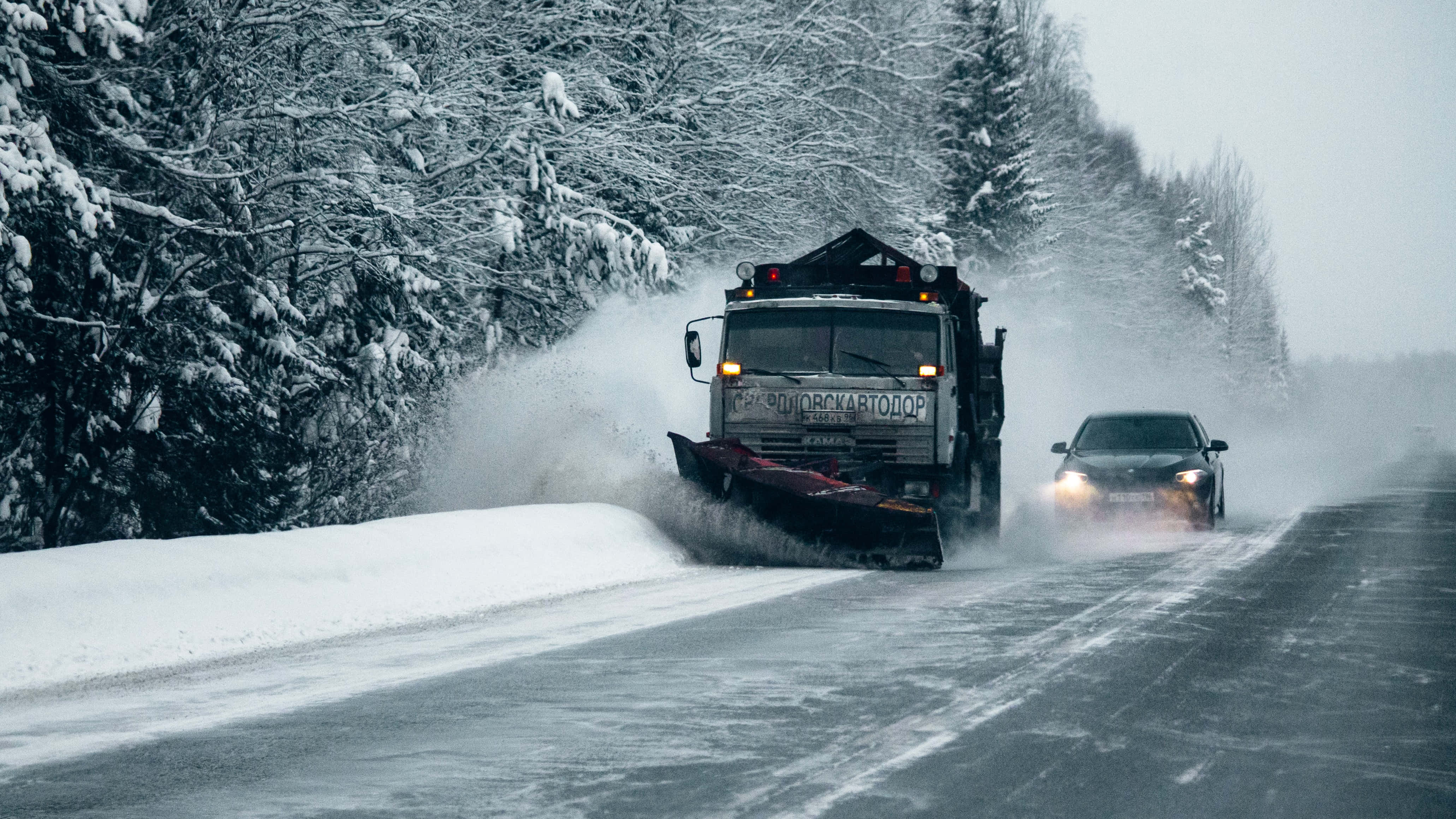 Snowplow 桌布 壁紙 — 一輛鏟雪車正在清理一條被雪覆蓋的道路，道路兩旁是積雪的樹木，一輛汽車在冬季風暴中跟在後面。