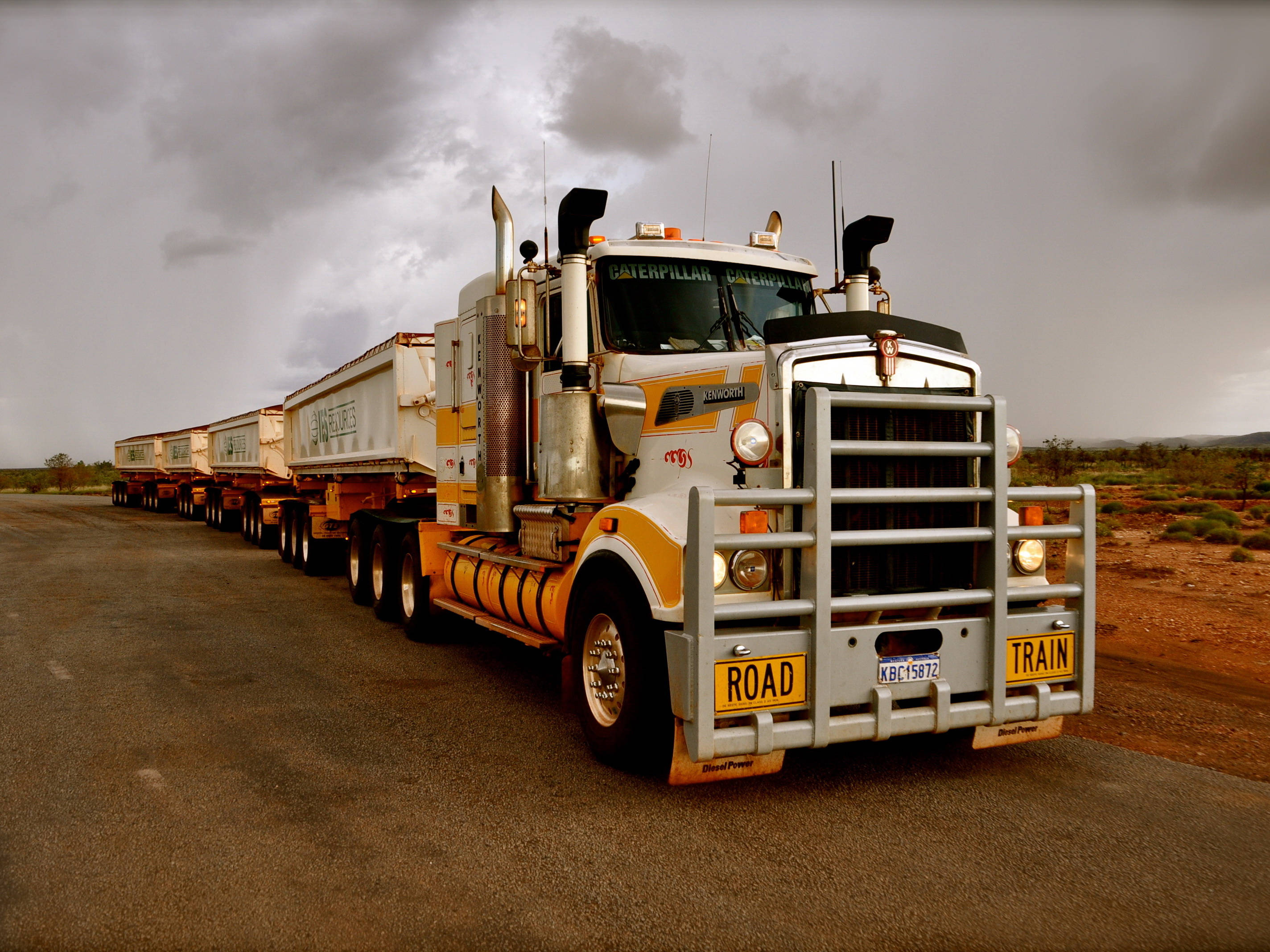 Trucks 桌布 壁紙 — 一輛長長的黃白色半掛卡車，保險桿上帶有「ROAD」和「TRAIN」標誌，在多雲的天空下行駛在一條荒涼的沙漠公路上。