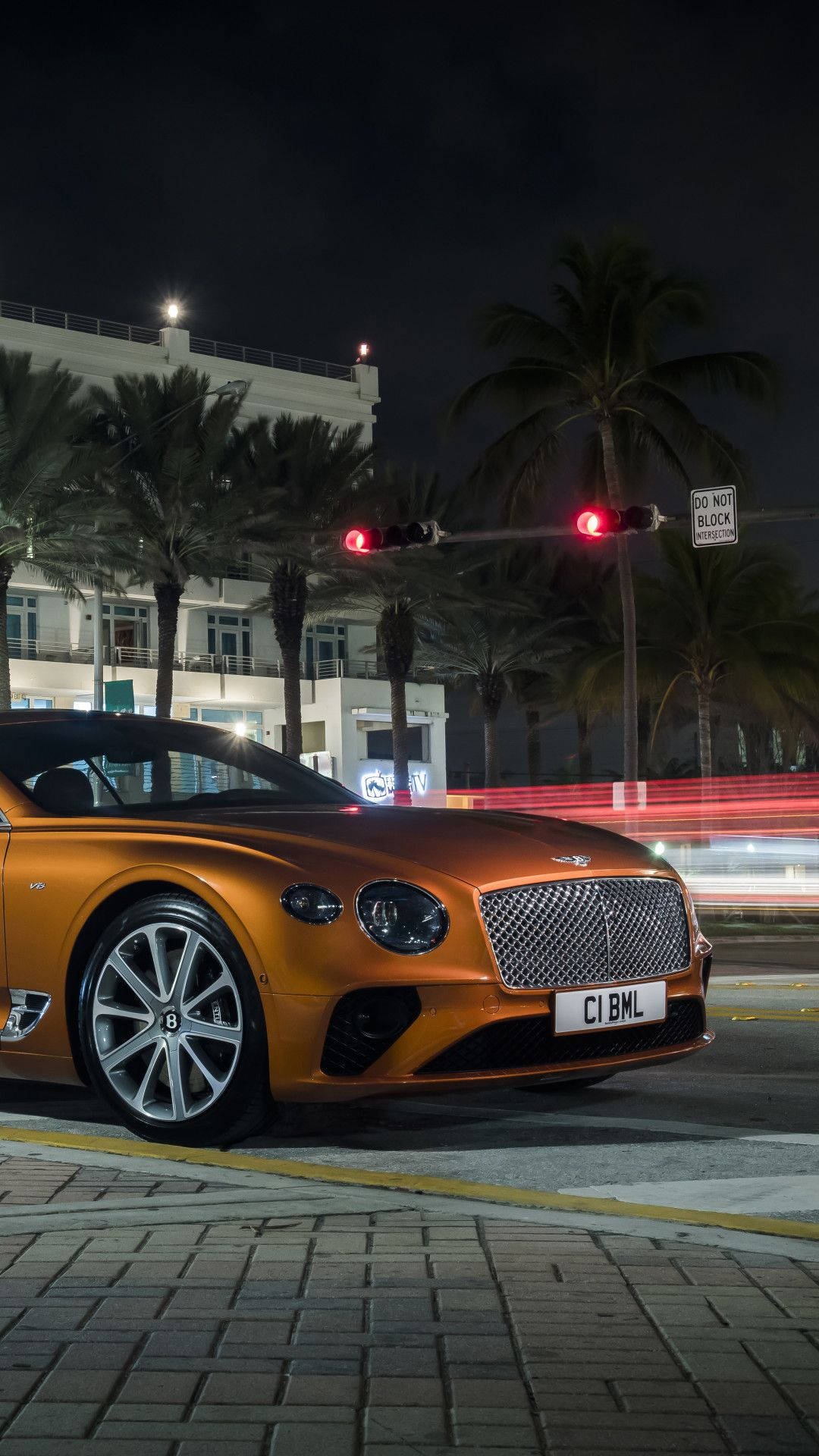 手機 桌布 壁紙 — 一輛閃亮的橙色 Bentley Continental GT 在夜間停泊在棕櫚樹下，背景有紅色交通燈閃爍。