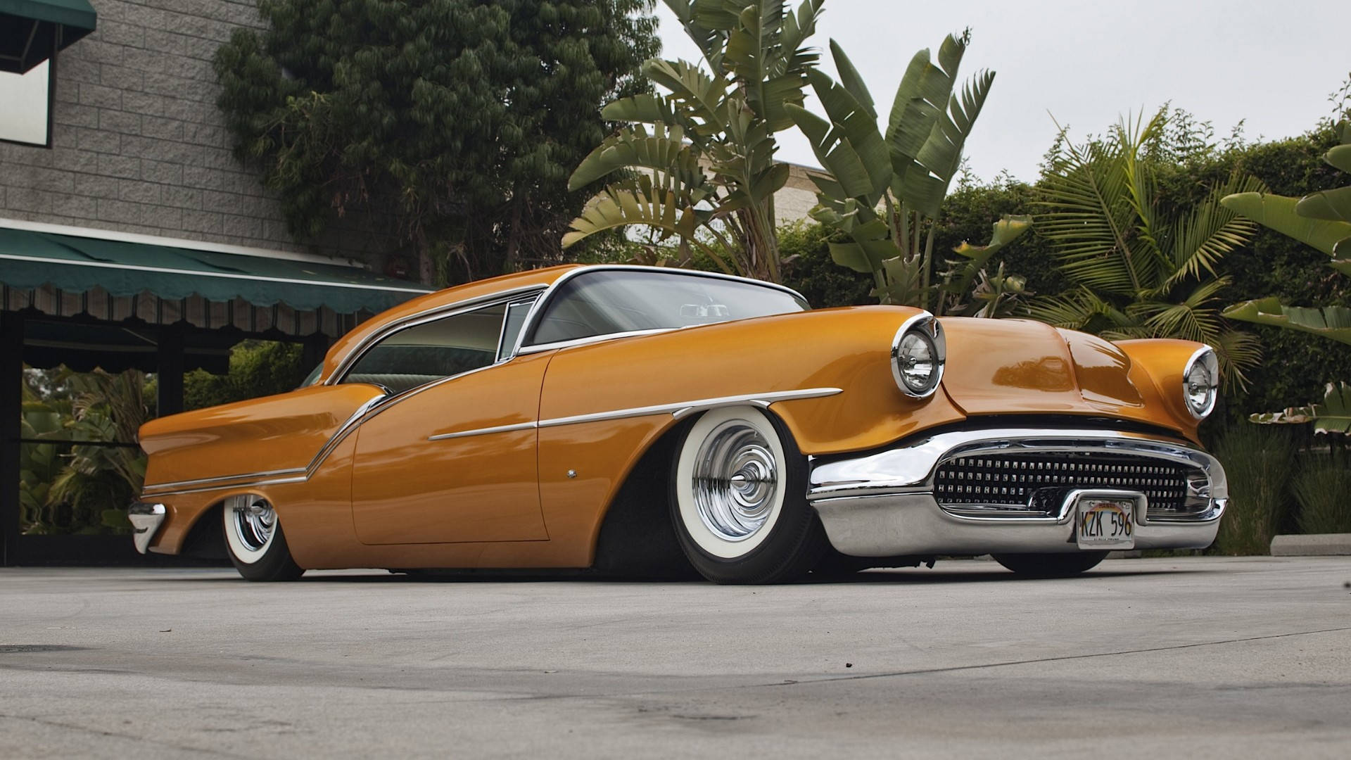 Lowrider 桌布 壁紙 — 一輛閃亮的橙色經典汽車，帶有鍍鉻細節，停在混凝土車道上，旁邊是茂密的綠色棕櫚樹。