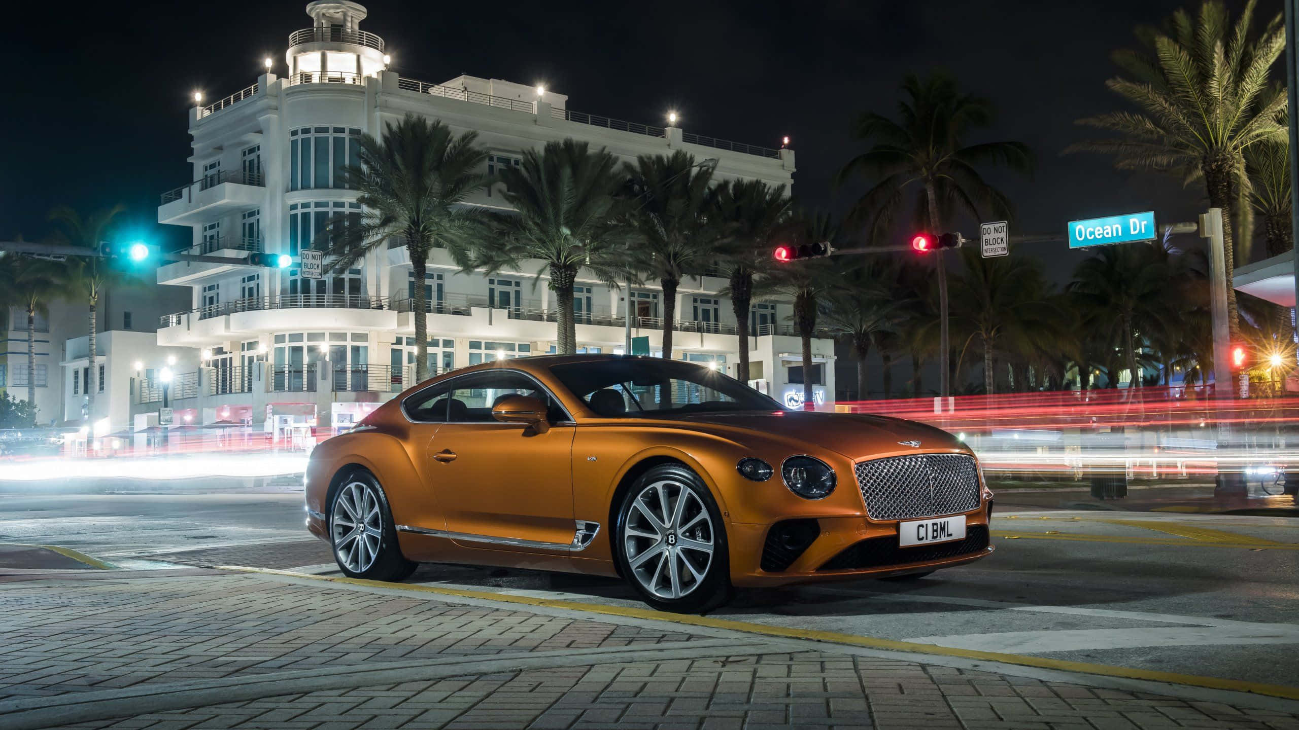 Best Bentley 桌布 壁紙 — 一輛閃亮的橙色賓利 Continental GT 在夜間停泊在海洋大道上，背景有棕櫚樹和光跡。