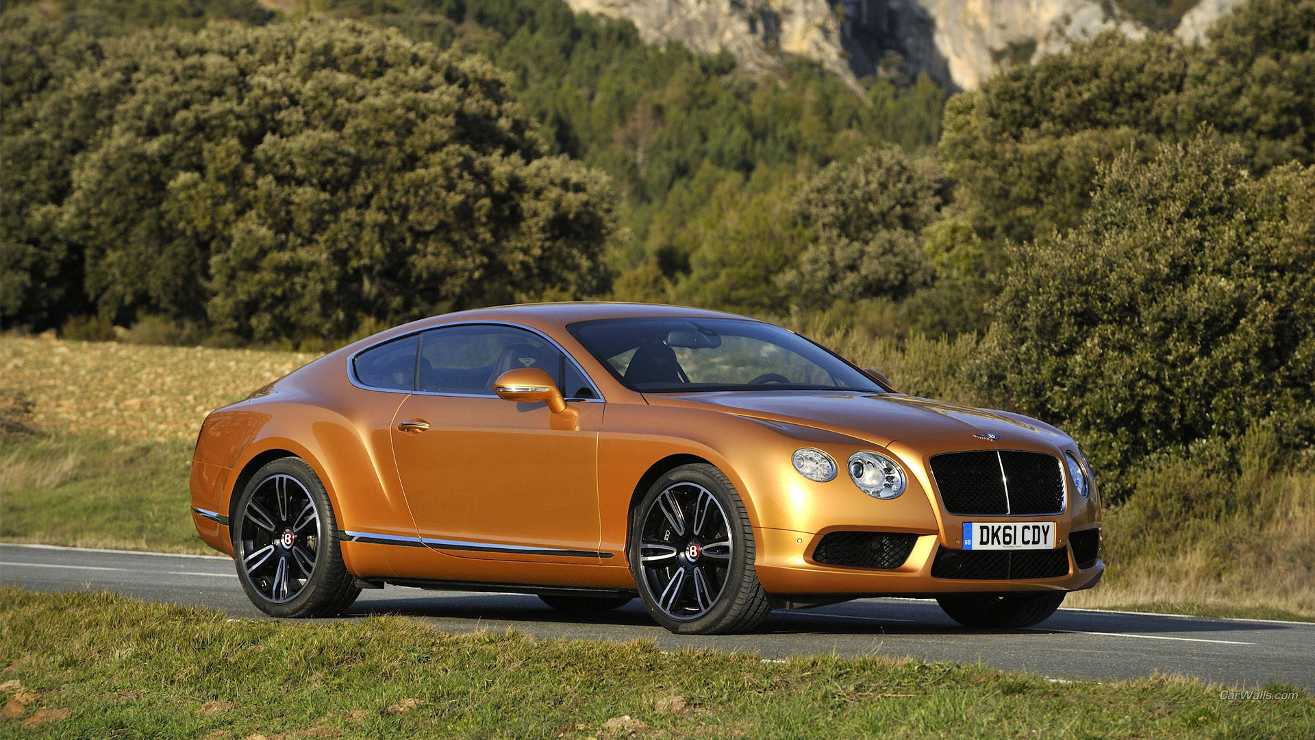 bentley HD 桌布 壁紙 — 一輛閃亮的橙色賓利 Continental GT 在明亮的日光下行駛在一條蜿蜒的鄉村道路上，兩旁是茂密的綠樹和岩石丘陵。