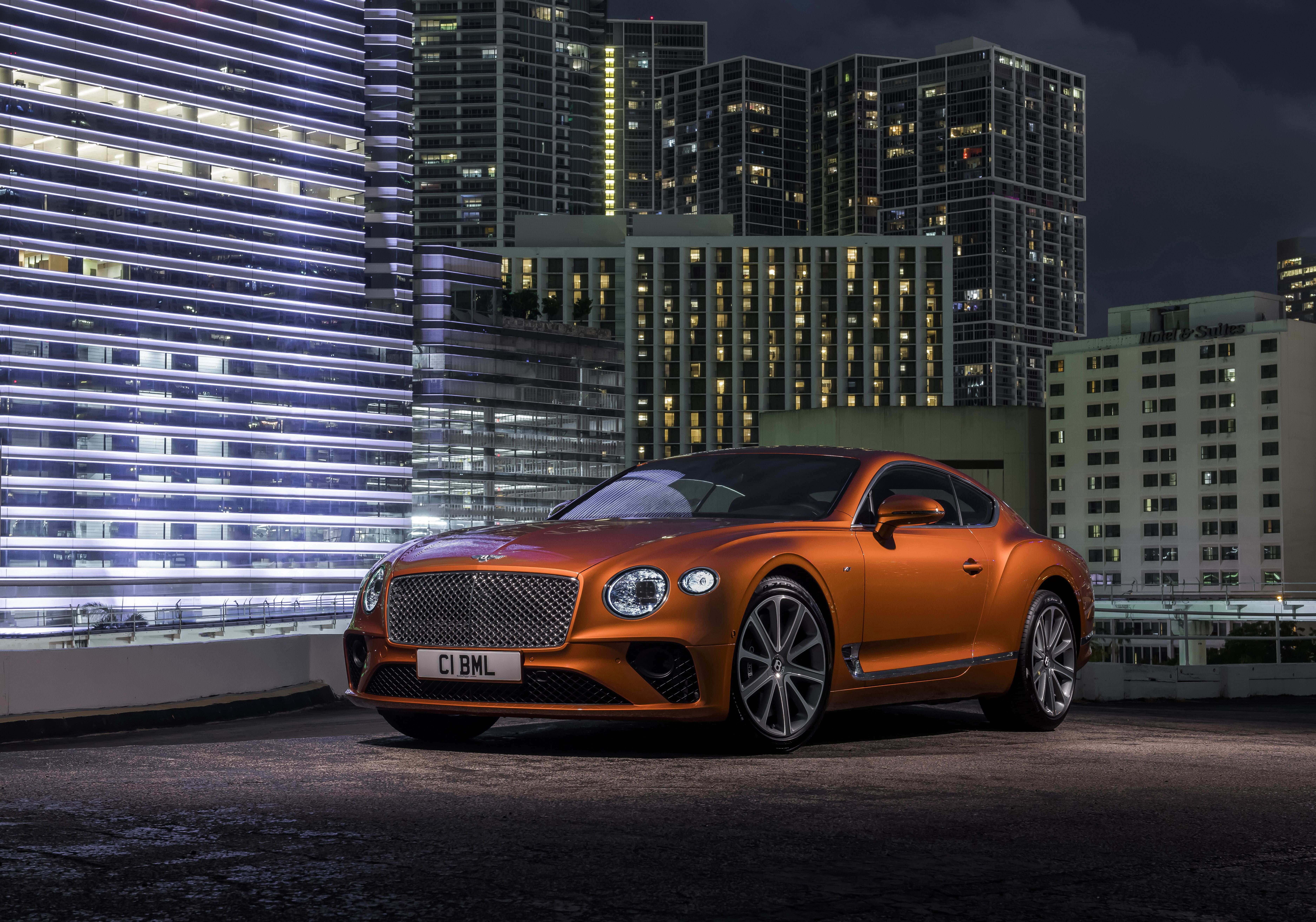Best Bentley 桌布 壁紙 — 一輛閃亮的橙色賓利 Continental GT 在夜間停泊在天台上，背後是燈火通明的摩天大樓。