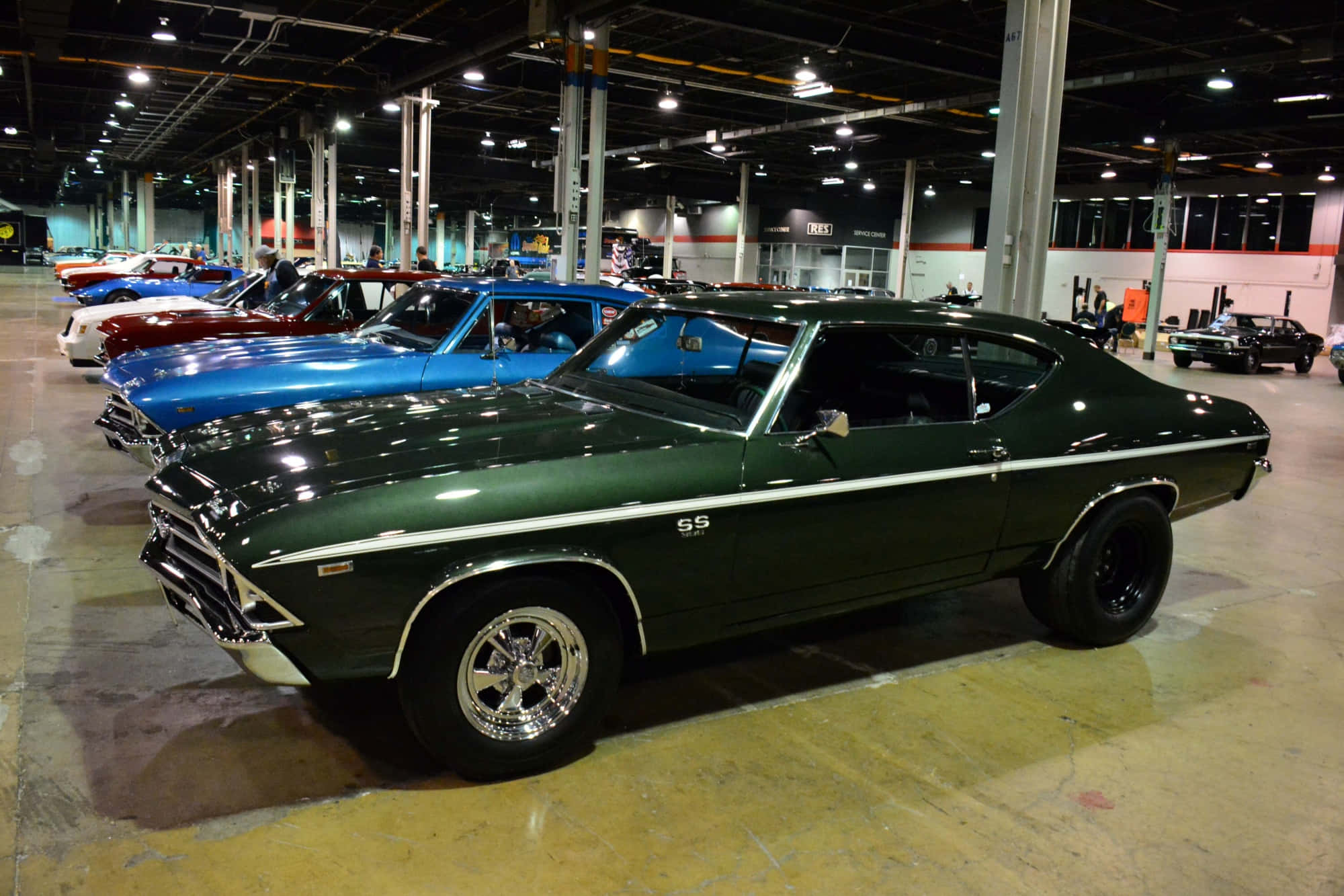 Muscle Cars 桌布 壁紙 — 一輛閃亮的深綠色經典肌肉車，帶有鍍鉻細節和黑色輪胎，停放在一個寬敞的室內汽車展廳，旁邊有其他復古車輛。