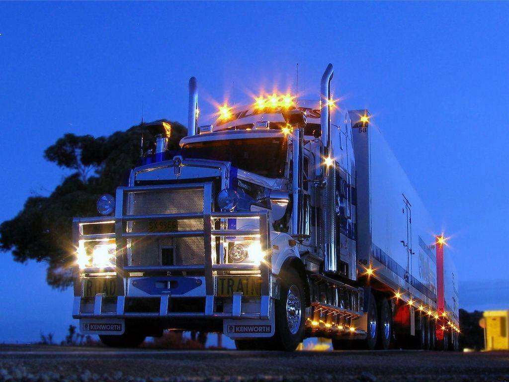 Kenworth 桌布 壁紙 — 一輛閃亮的白色Kenworth半掛卡車，明亮的車頭燈和琥珀色警告燈照亮了黑暗樹木旁的暮色道路。