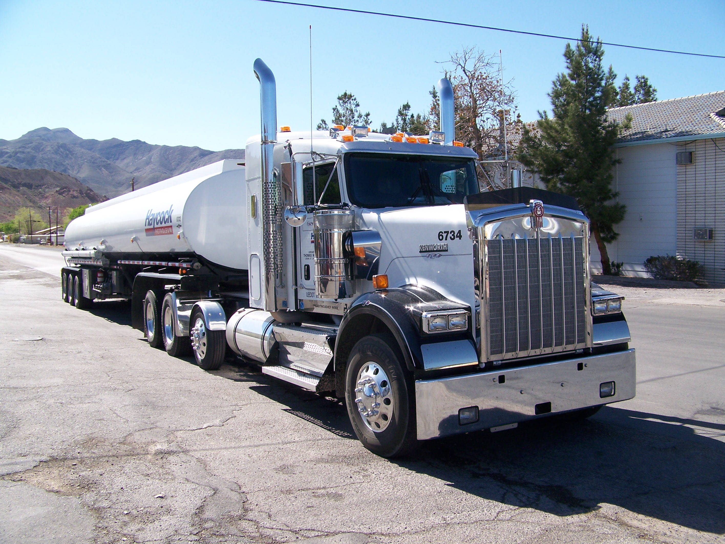 Kenworth W900 桌布 壁紙 — 一輛閃亮的白色Kenworth油罐車，帶有鍍鉻裝飾，停在裂開的瀝青路上，背景是晴朗的藍天和山脈。