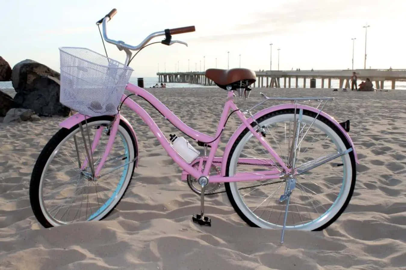 Beach Cruiser 桌布 壁紙 — 一輛閃亮的粉紅色沙灘巡航自行車，配有鐵絲籃，停放在日落時分碼頭附近的沙灘上。