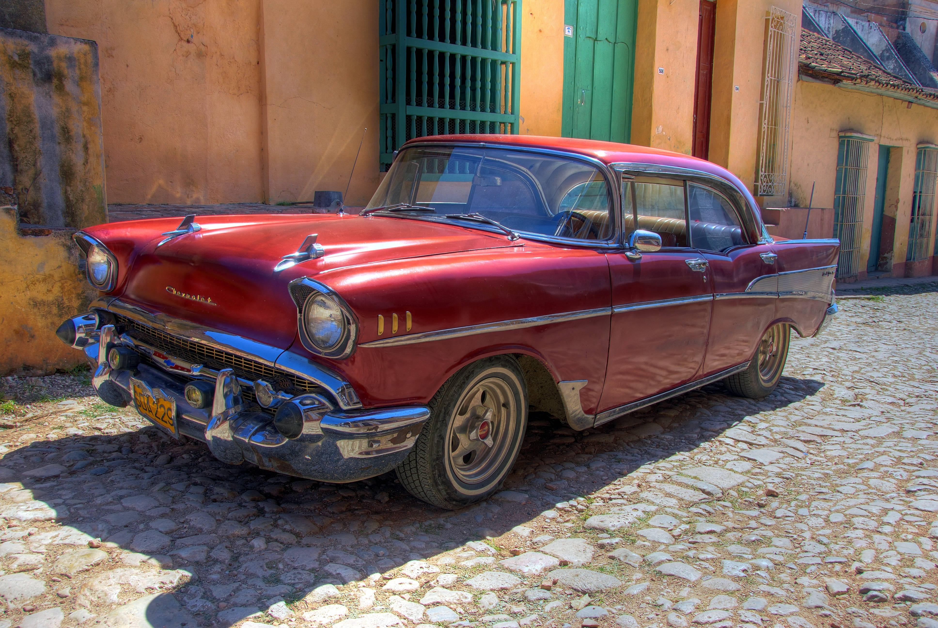 Cuba 桌布 壁紙 — 一輛閃亮的紅色1957年雪佛蘭Bel Air在明亮的陽光下，在兩旁是粉彩殖民建築的風化鵝卵石街道上閃閃發光。