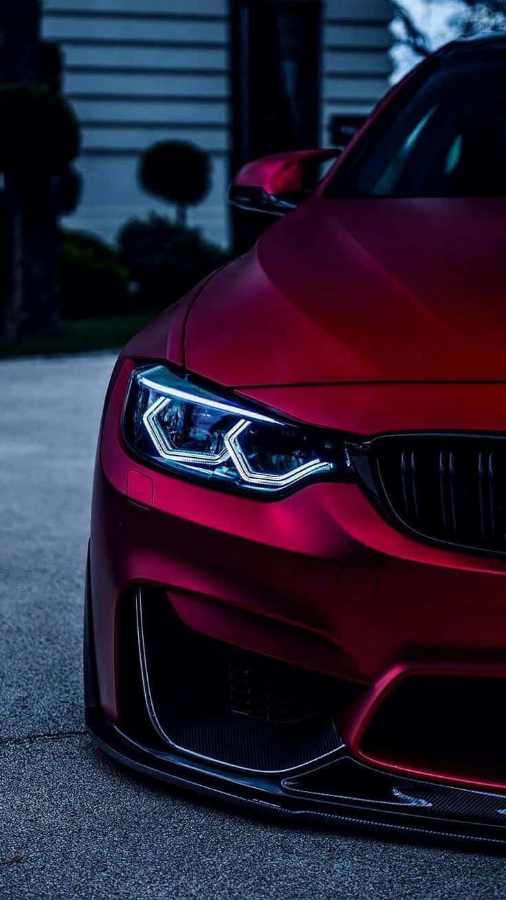 Bmw iPhone 桌布 壁紙 — 一輛閃亮的紅色BMW跑車，車頭燈發光，停泊在暮色中的車道上。