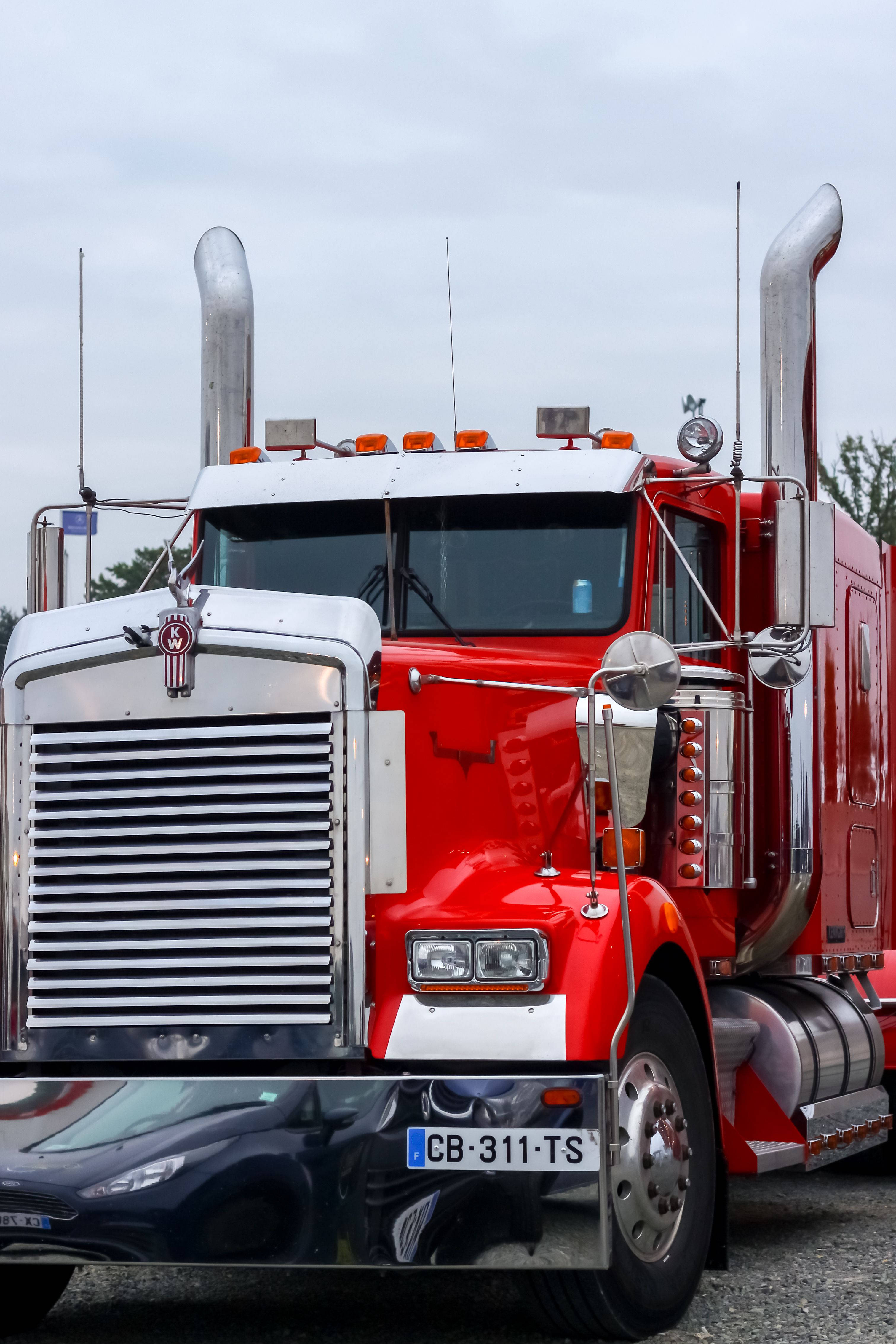 Kenworth 桌布 壁紙 — 一輛閃亮的紅色Kenworth半掛卡車，帶有鍍鉻裝飾和雙排氣管，停在陰天之下。