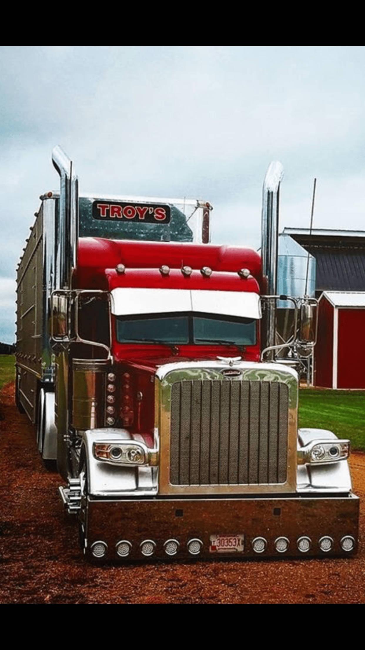 Peterbilt 桌布 壁紙 — 一輛閃亮的紅色半掛卡車，帶有鍍鉻裝飾和'TROY'S'標誌，停泊在紅色穀倉附近的泥土上，天空多雲。