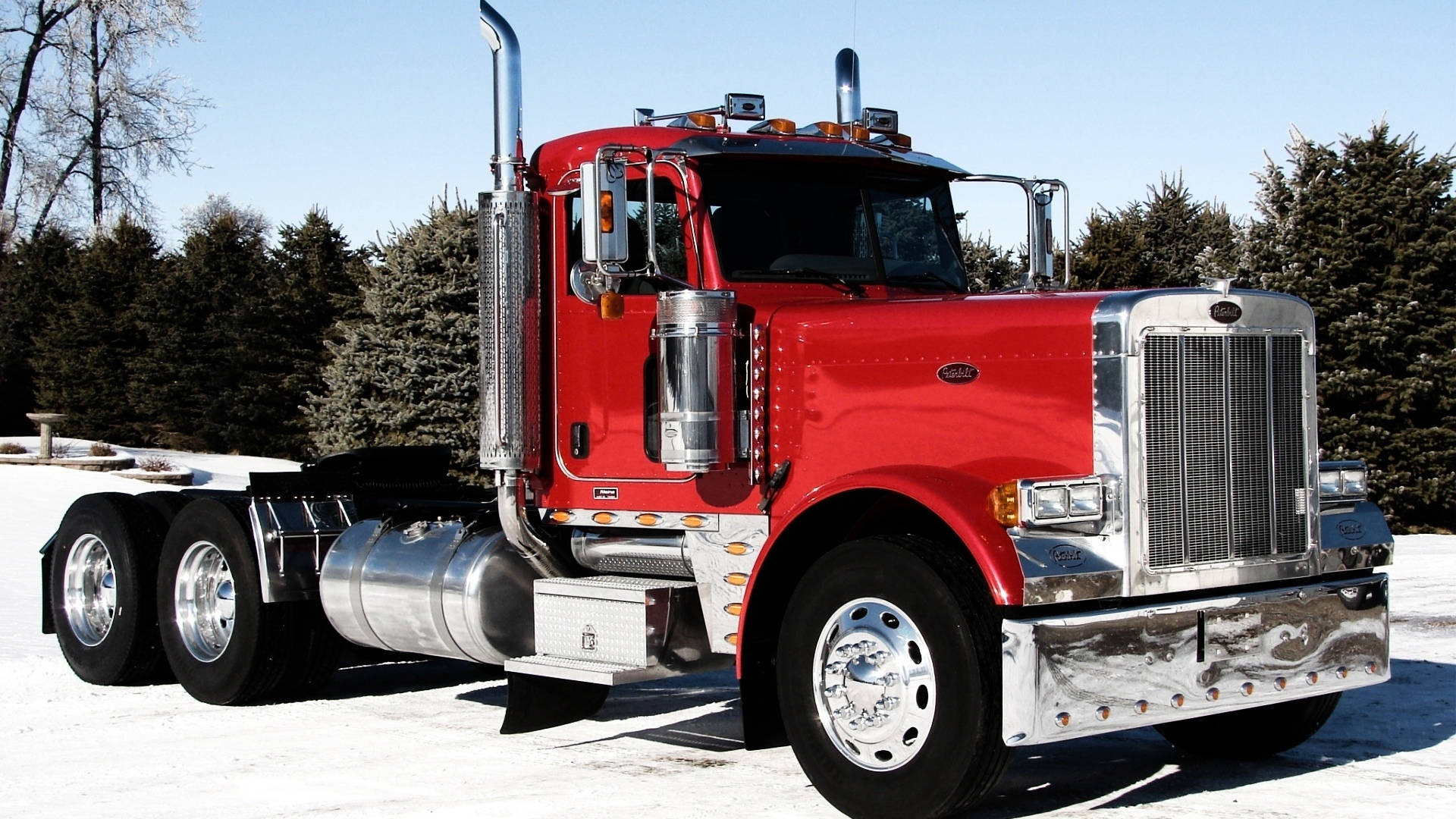 Peterbilt 桌布 壁紙 — 一輛閃亮的紅色半掛卡車帶有鍍鉻裝飾，停在雪地上，背景是晴朗的藍天和常綠樹。