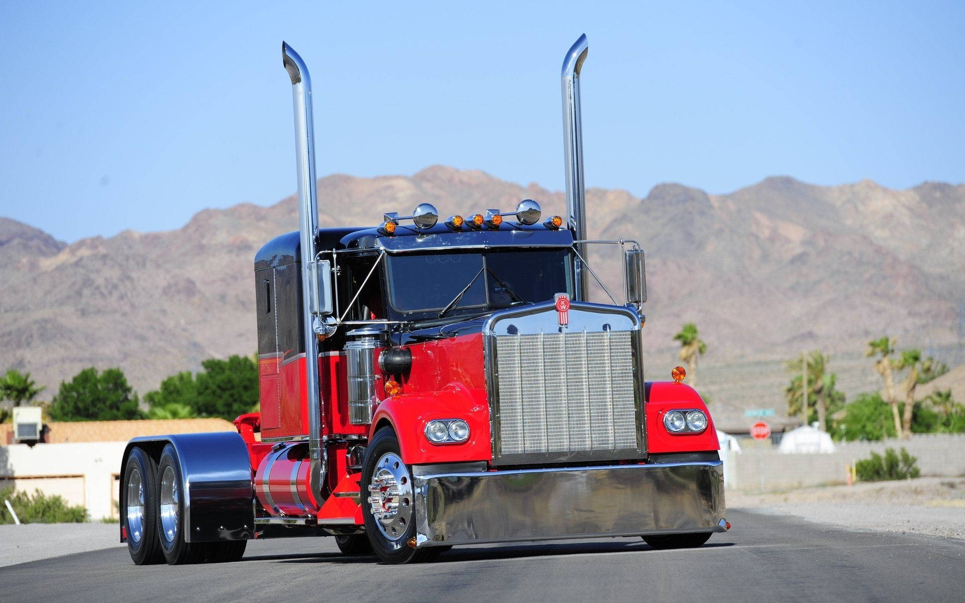 Kenworth 桌布 壁紙 — 一輛閃亮的紅色和鍍鉻半掛卡車，帶有高高的排氣管，在晴朗的藍天下行駛在沙漠公路上，背景是山脈。