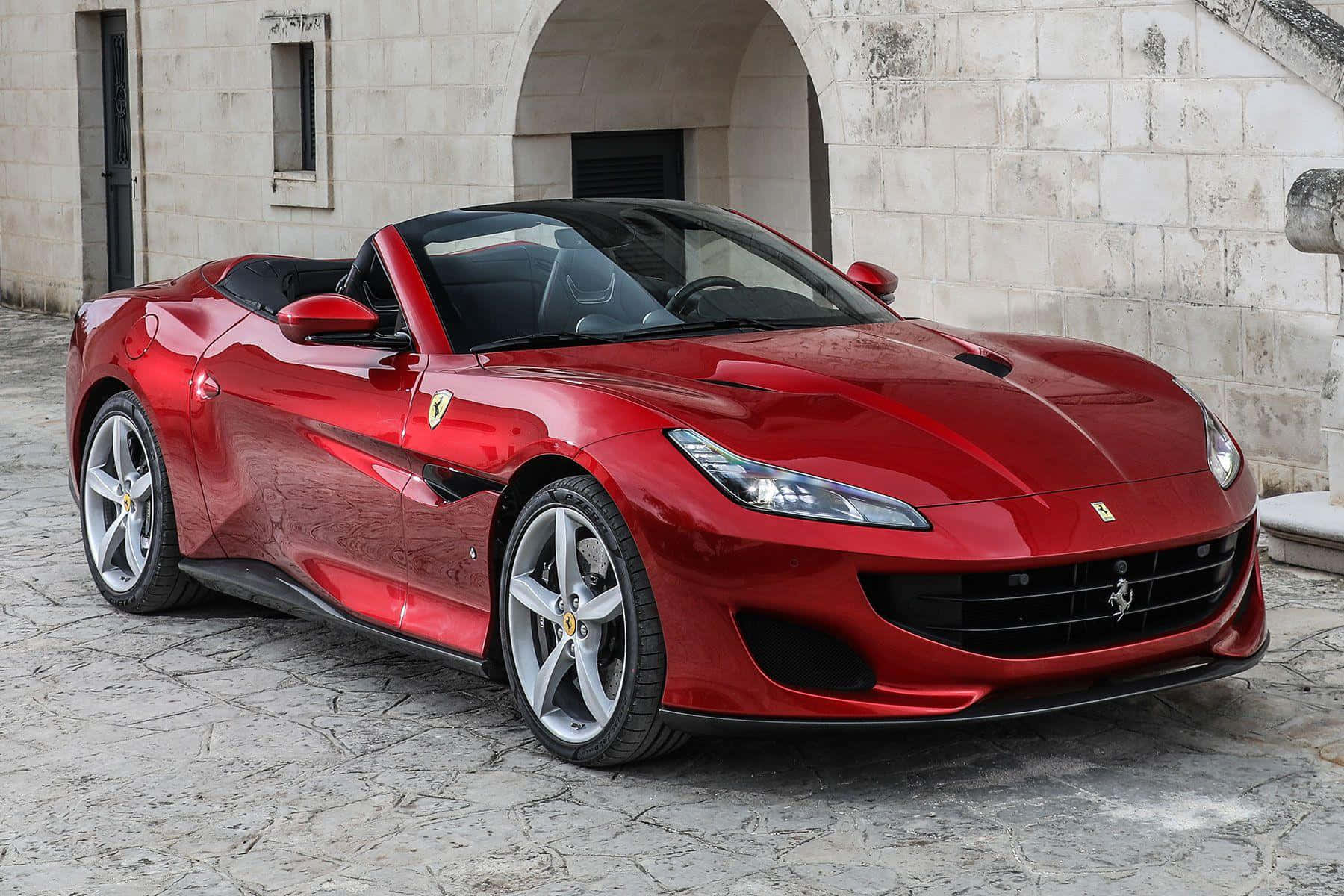 Ferrari Portofino 桌布 壁紙 — 一輛閃亮的紅色法拉利Portofino敞篷車停放在陽光明媚的石頭庭院中，旁邊是一座歷史悠久的石頭建築。