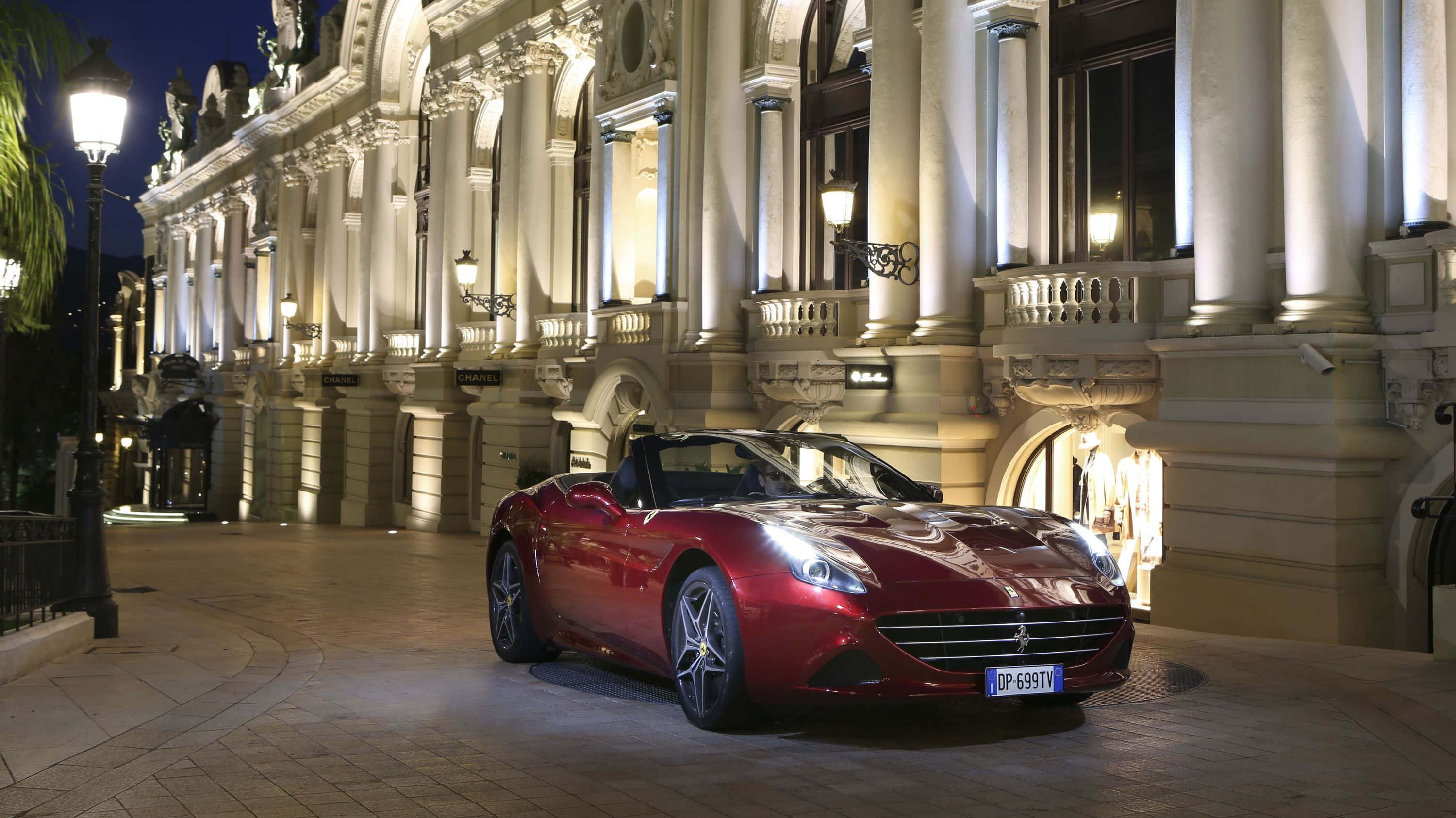 Ferrari California T 桌布 壁紙 — 一輛閃亮的紅色法拉利敞篷車在夜間停在一棟裝飾華麗、燈光璀璨的歐式建築前。