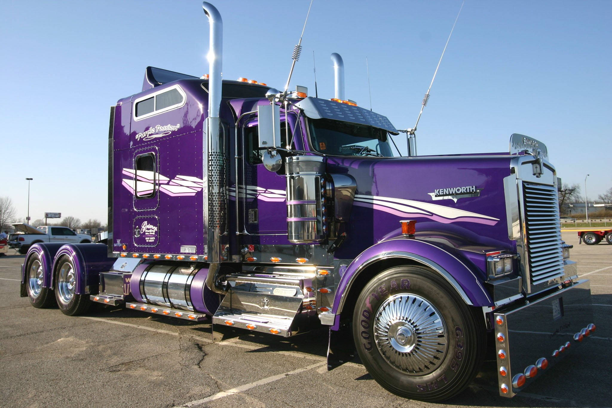 Kenworth W900 桌布 壁紙 — 一輛閃亮的紫色Kenworth半掛卡車，帶有鍍鉻裝飾和定制貼紙，停在陽光明媚的停車場。
