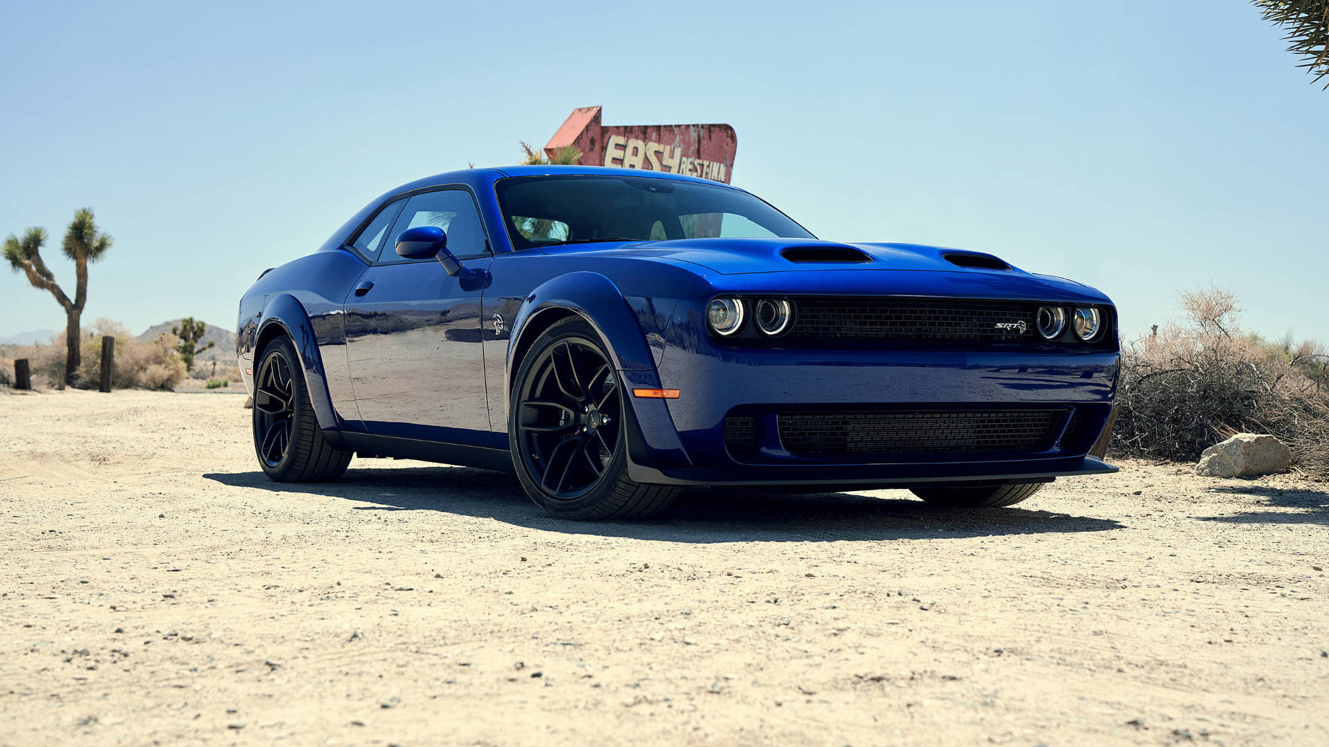沙漠 桌布 壁紙 — 一輛閃亮的藍色 Dodge Challenger 肌肉車停泊在塵土飛揚的沙漠道路上，背景是晴朗的藍天和一個褪色的路邊標誌。