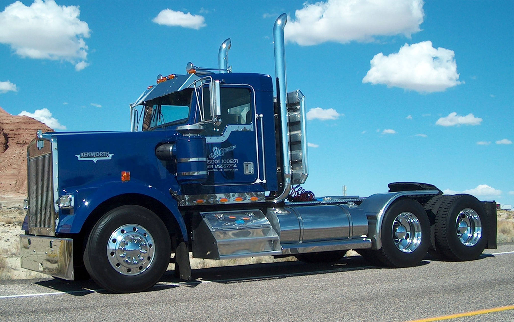 Kenworth W900 桌布 壁紙 — 一輛閃亮的藍色 Kenworth 半掛卡車，帶有鍍鉻裝飾，停在沙漠公路上，天空湛藍，白雲朵朵。