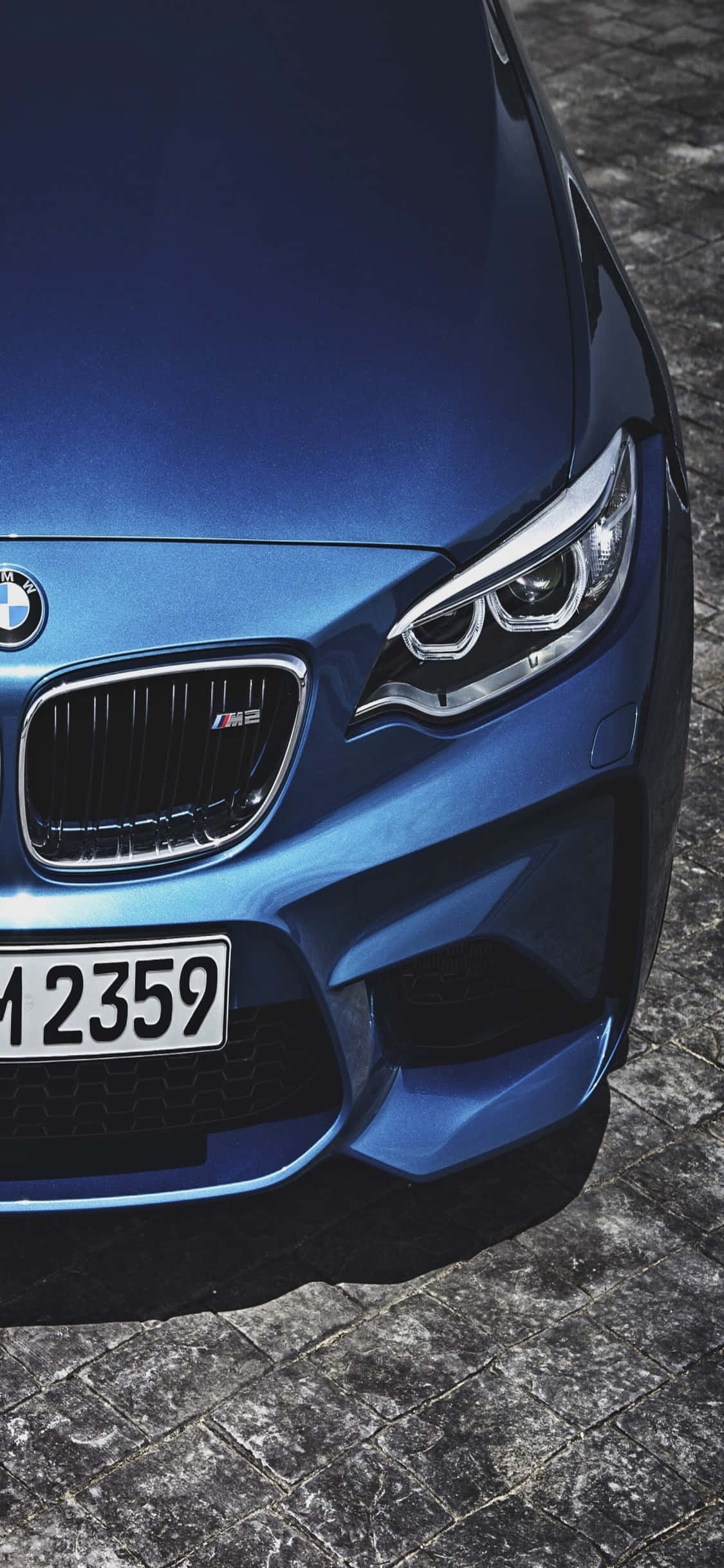 iPhone X Bmw 桌布 壁紙 — 一輛閃亮的藍色BMW M2跑車停放在有紋理的鵝卵石街道上的特寫。