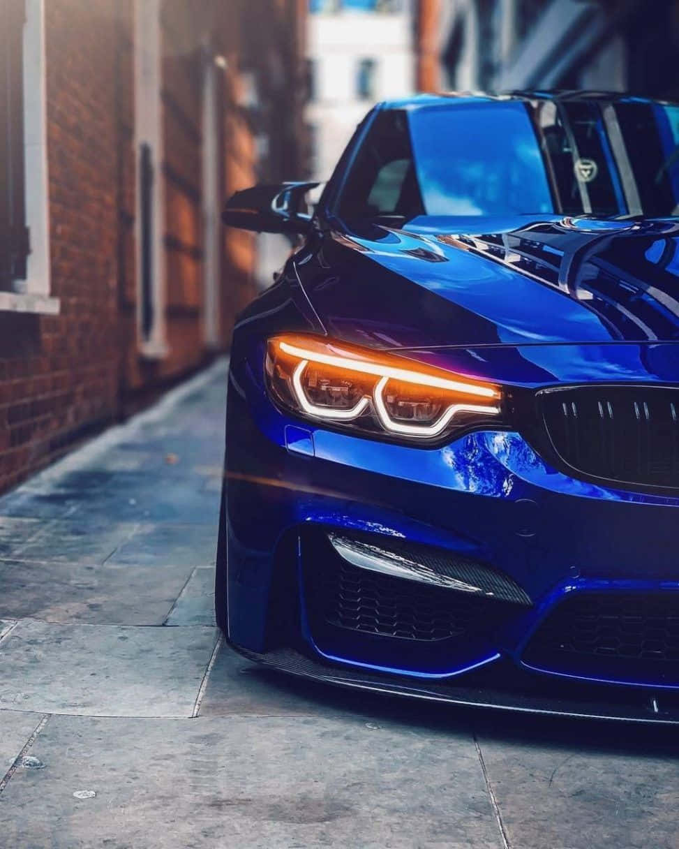 BMW 動圖 桌布 壁紙 — 一輛閃亮的藍色BMW跑車，亮著橙色車頭燈，停在一條狹窄的磚砌城市小巷中。