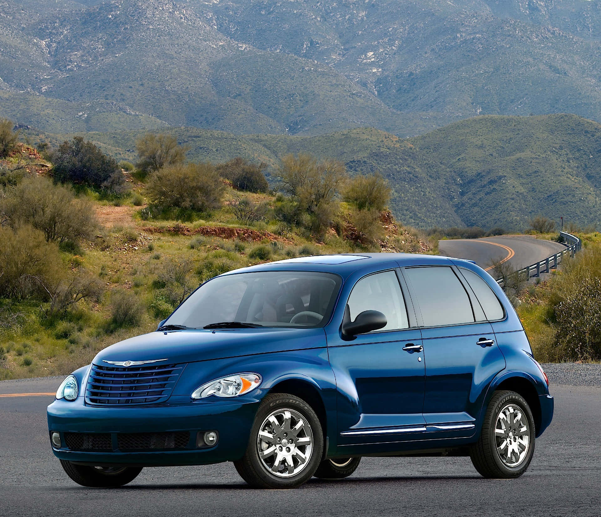Chrysler Pt Cruiser 桌布 壁紙 — 一輛閃亮的藍色PT Cruiser在明亮的日光下行駛在一條蜿蜒的山路上，穿過綠色的山丘。