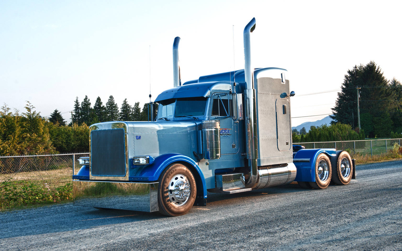 Peterbilt 桌布 壁紙 — 一輛閃亮的藍色半掛卡車，帶有鍍鉻裝飾，停在礫石上，旁邊是鏈條圍欄，天空明亮。