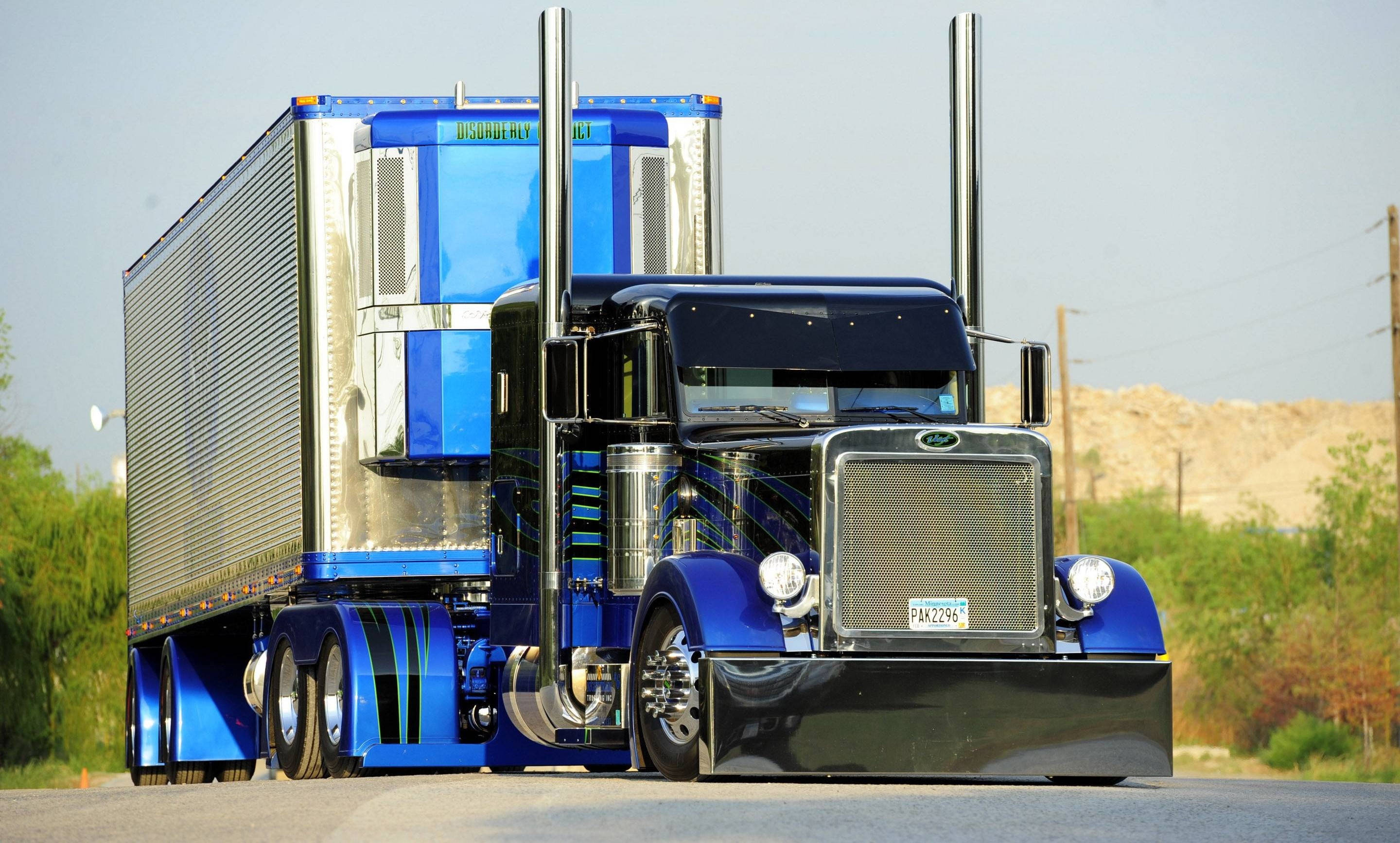Peterbilt 桌布 壁紙 — 一輛閃亮的藍色半掛卡車，帶有鍍鉻裝飾和高排氣管，在明亮的日光下主宰著沙漠公路。