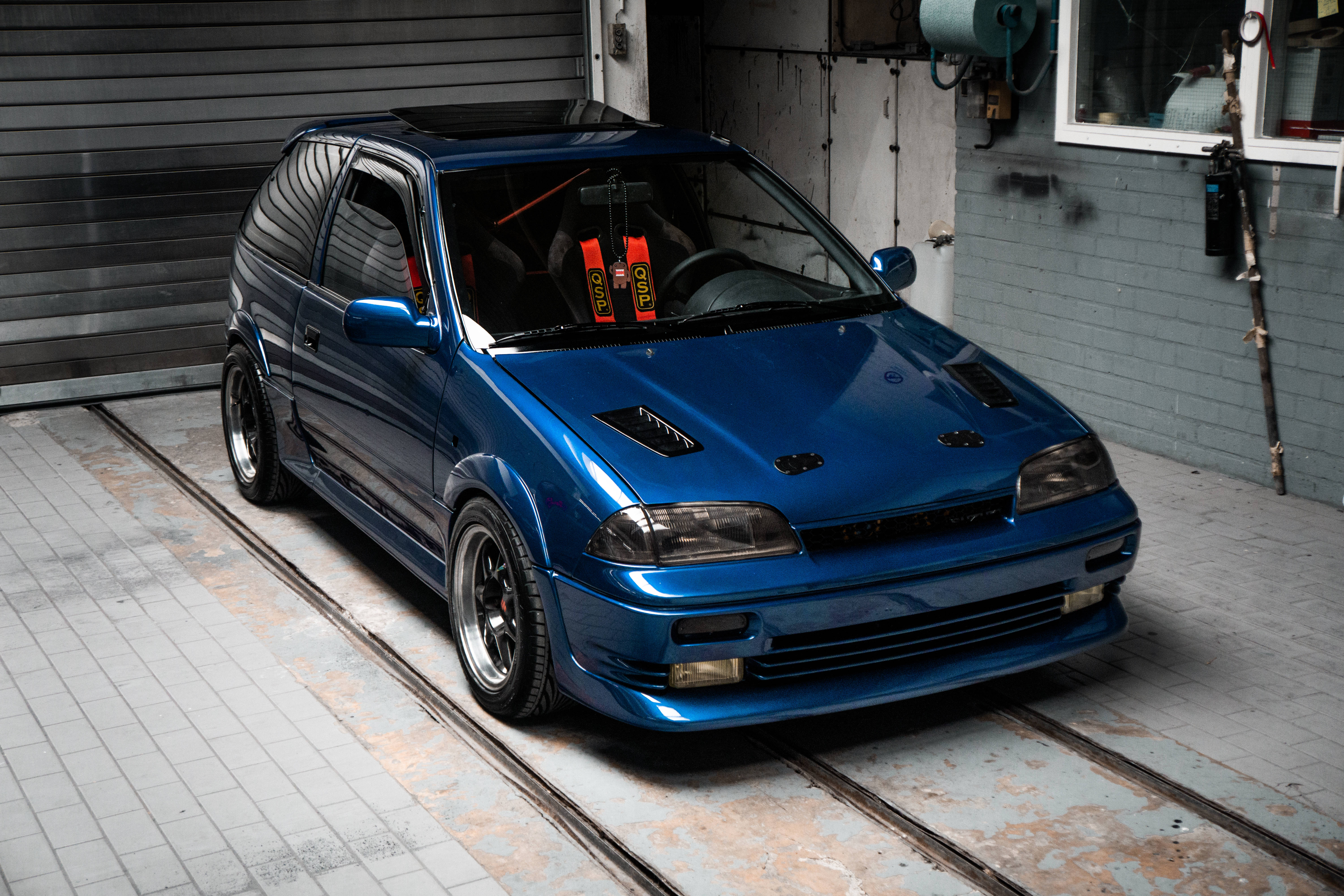 Suzuki 桌布 壁紙 — 一輛閃亮的藍色改裝掀背車停在一個有裸露軌道和工業牆壁的骯髒車庫中。