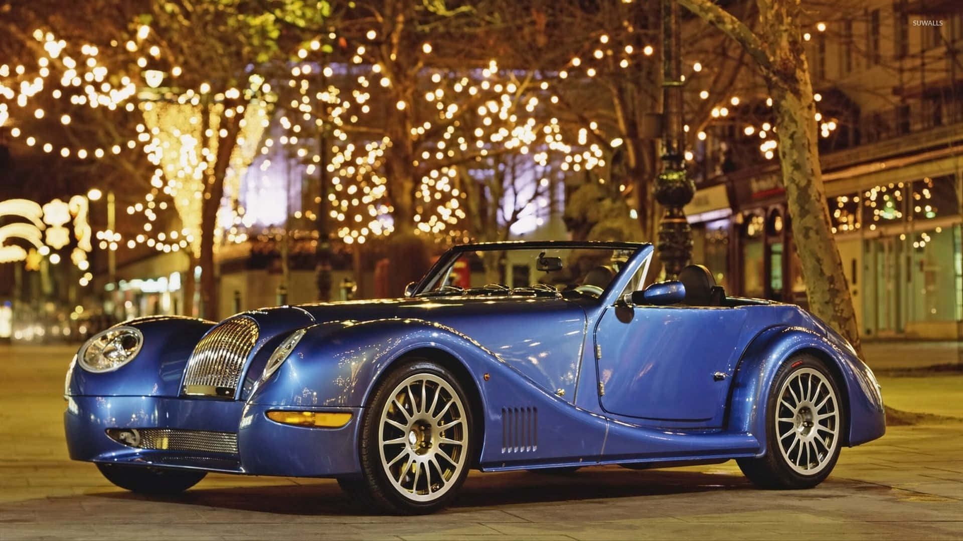 Morgan 桌布 壁紙 — 一輛閃亮的藍色敞篷跑車在夜間節日城市街道的溫暖燈串下閃耀。