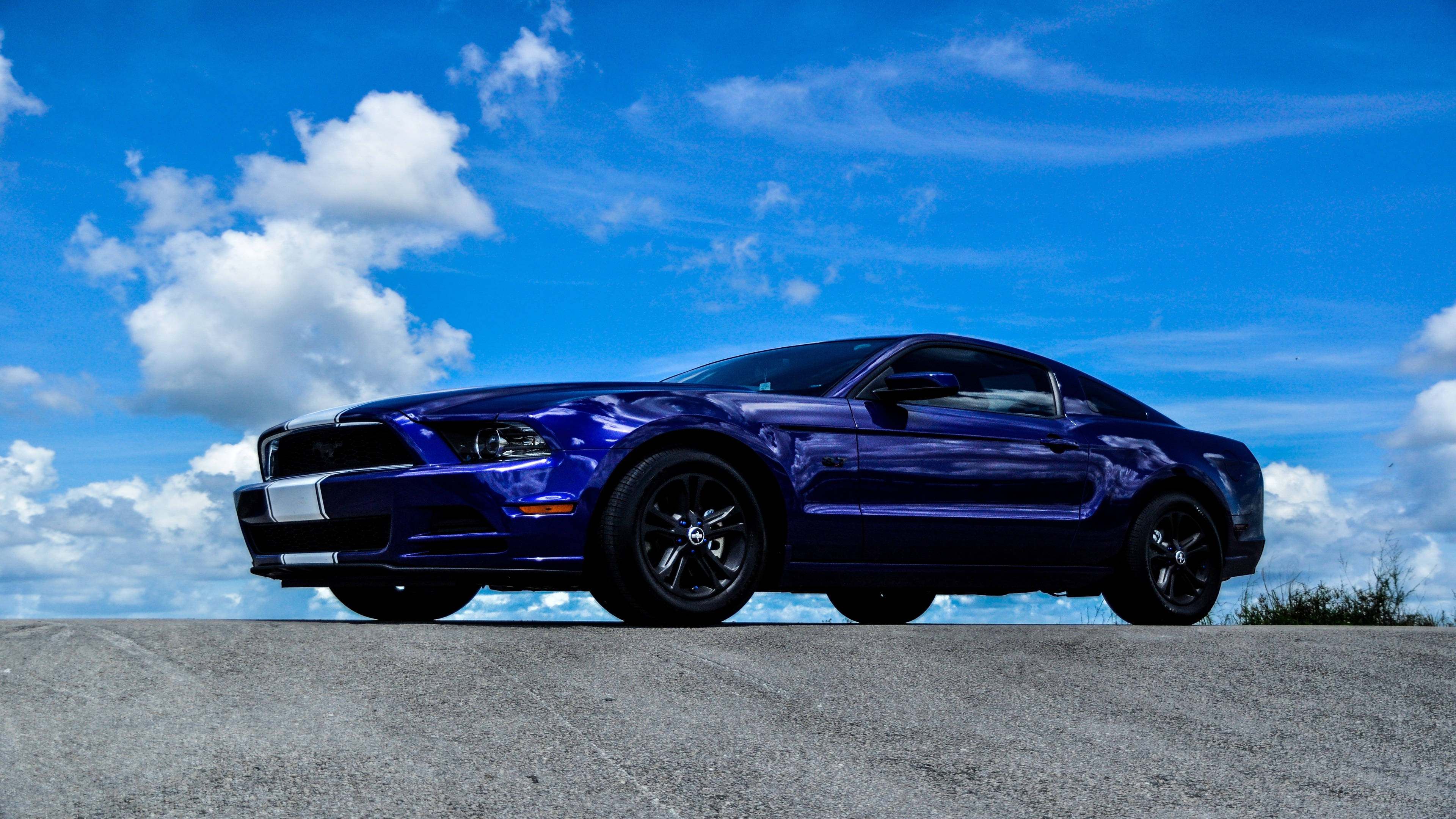 mustang HD 桌布 壁紙 — 一輛閃亮的藍色福特野馬跑車停在混凝土表面上，天空湛藍，白雲朵朵。