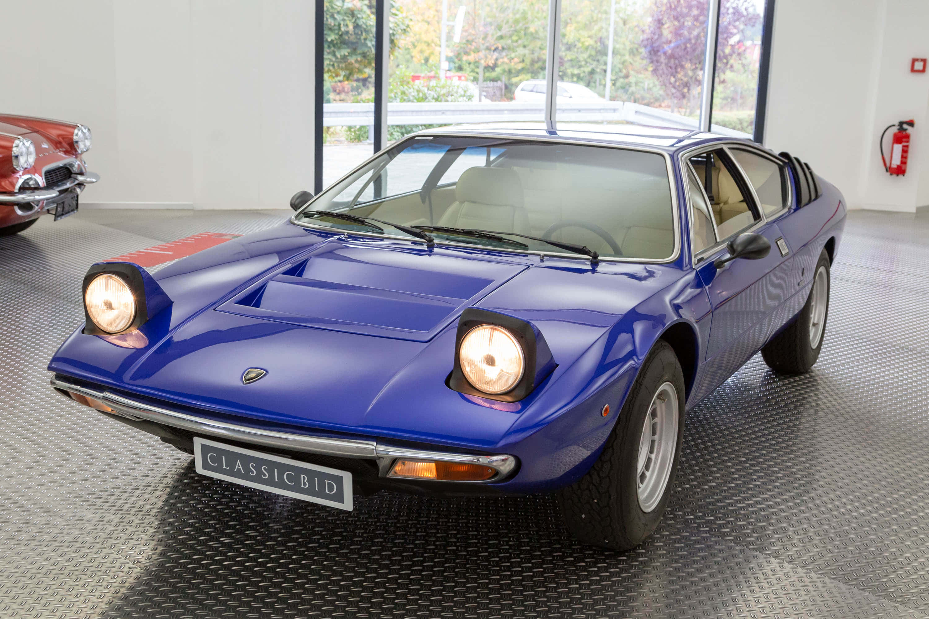 Lamborghini Urraco 桌布 壁紙 — 一輛閃亮的藍色經典藍寶堅尼Countach跑車在現代展廳的明亮燈光下閃耀，背景可見一輛紅色汽車。
