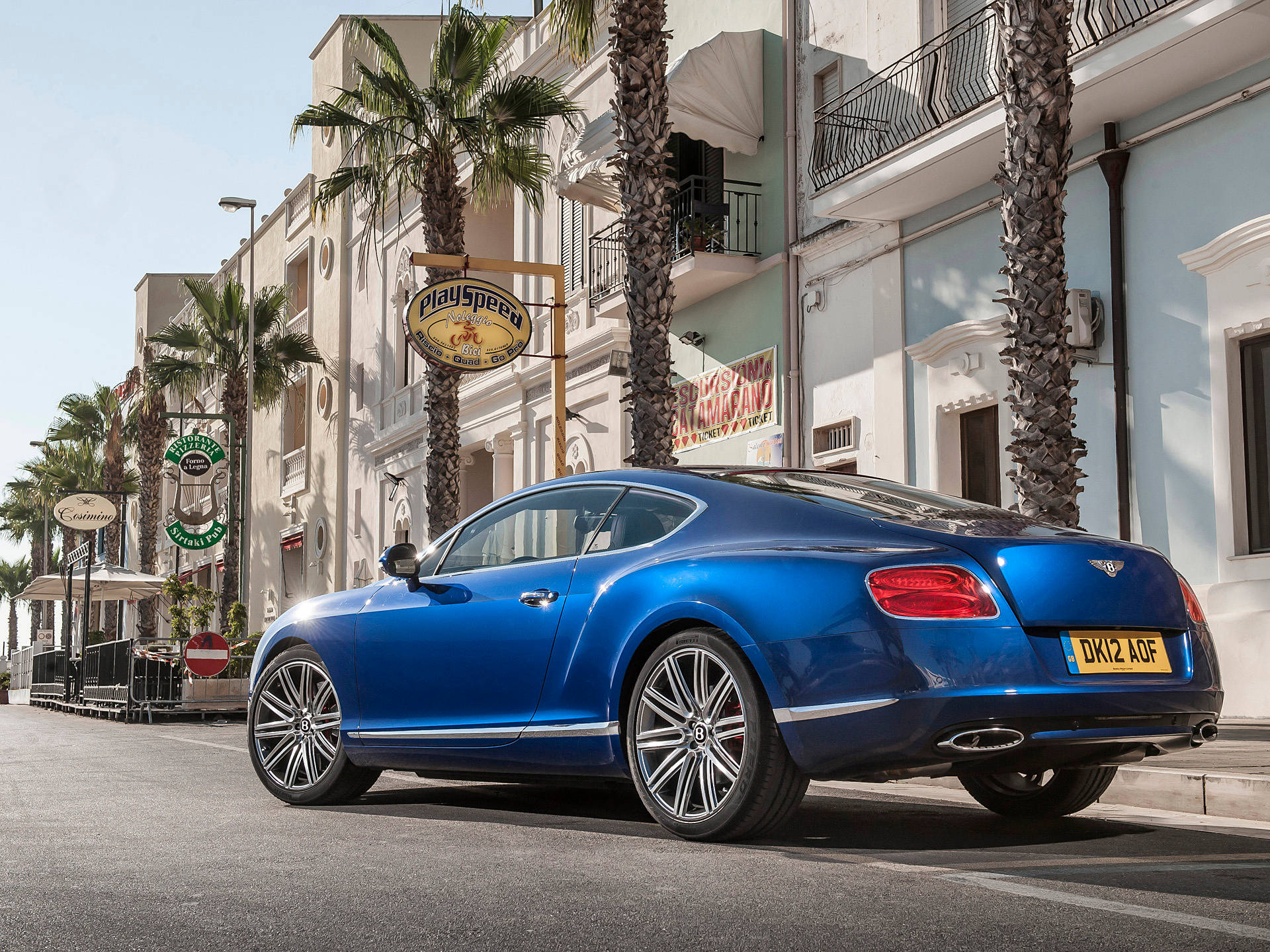 bentley HD 桌布 壁紙 — 一輛閃亮的藍色賓利 Continental GT 停泊在棕櫚樹成蔭的地中海街道上，兩旁是粉彩建築和明亮的陽光。