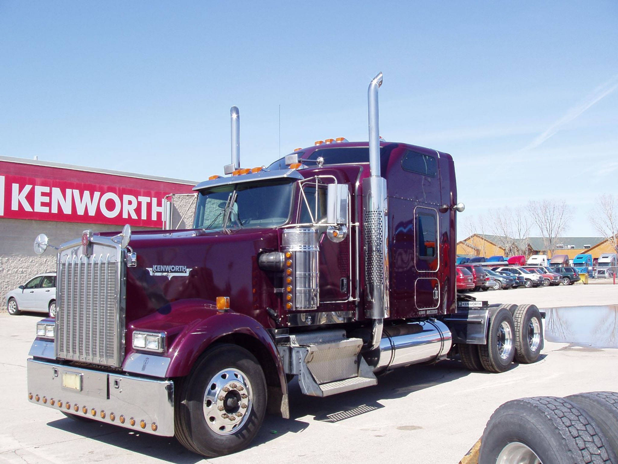 Kenworth W900 桌布 壁紙 — 一輛閃亮的酒紅色Kenworth半掛卡車帶有鍍鉻裝飾，停在陽光明媚的Kenworth經銷商大樓旁。