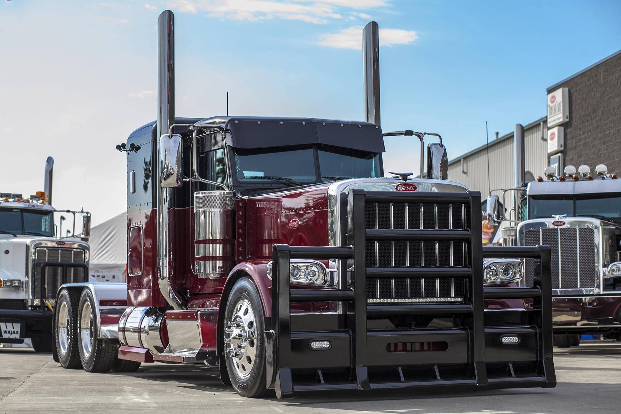 Peterbilt 桌布 壁紙 — 一輛閃亮的酒紅色Peterbilt半掛卡車，帶有鍍鉻裝飾和高排氣管，在陽光明媚的工業場地中顯眼地停放。