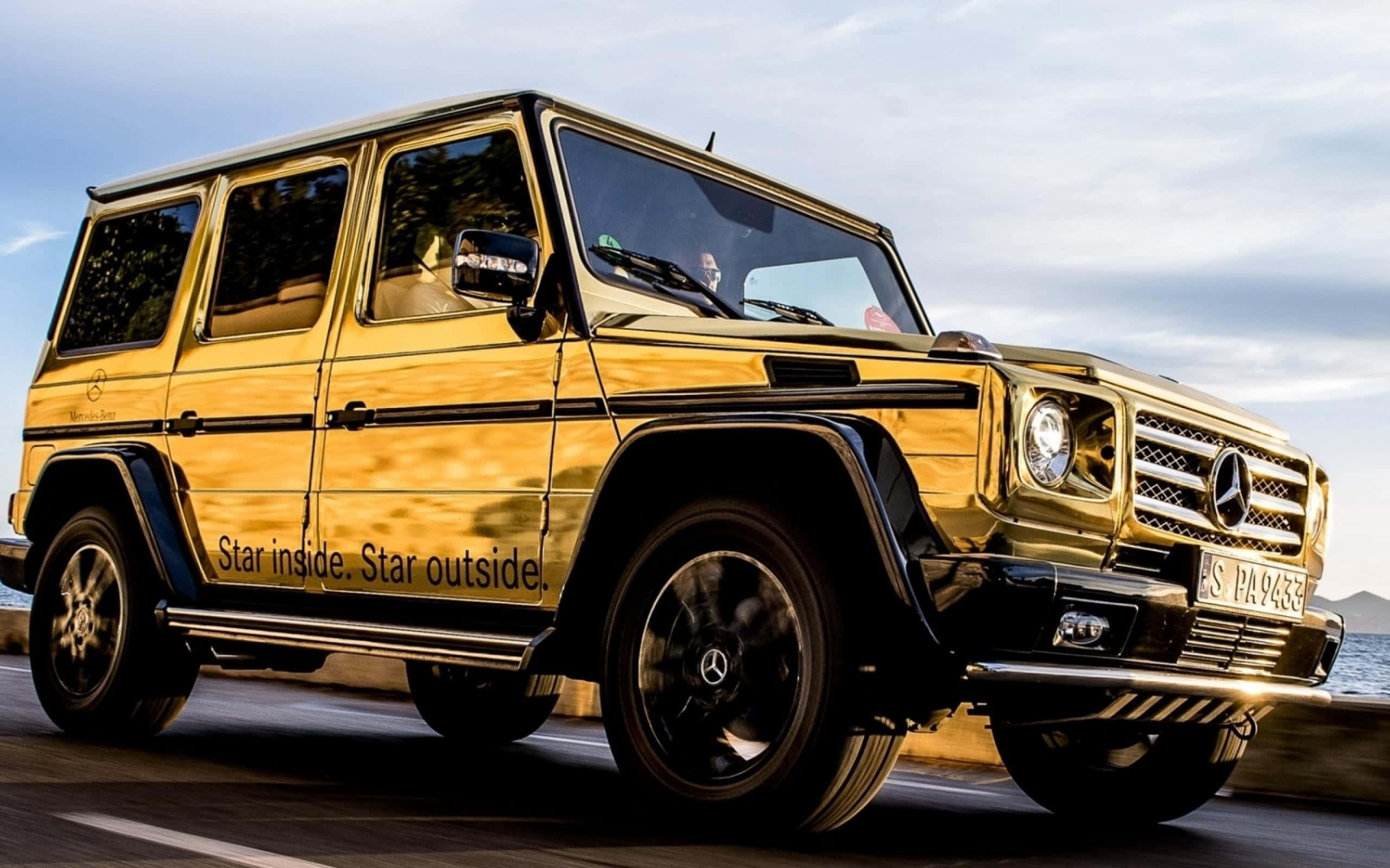 Gold Cars 桌布 壁紙 — 一輛閃亮的金色 Mercedes-Benz G-Class SUV 沿著海岸公路行駛，在陰天天空下欣賞海景。