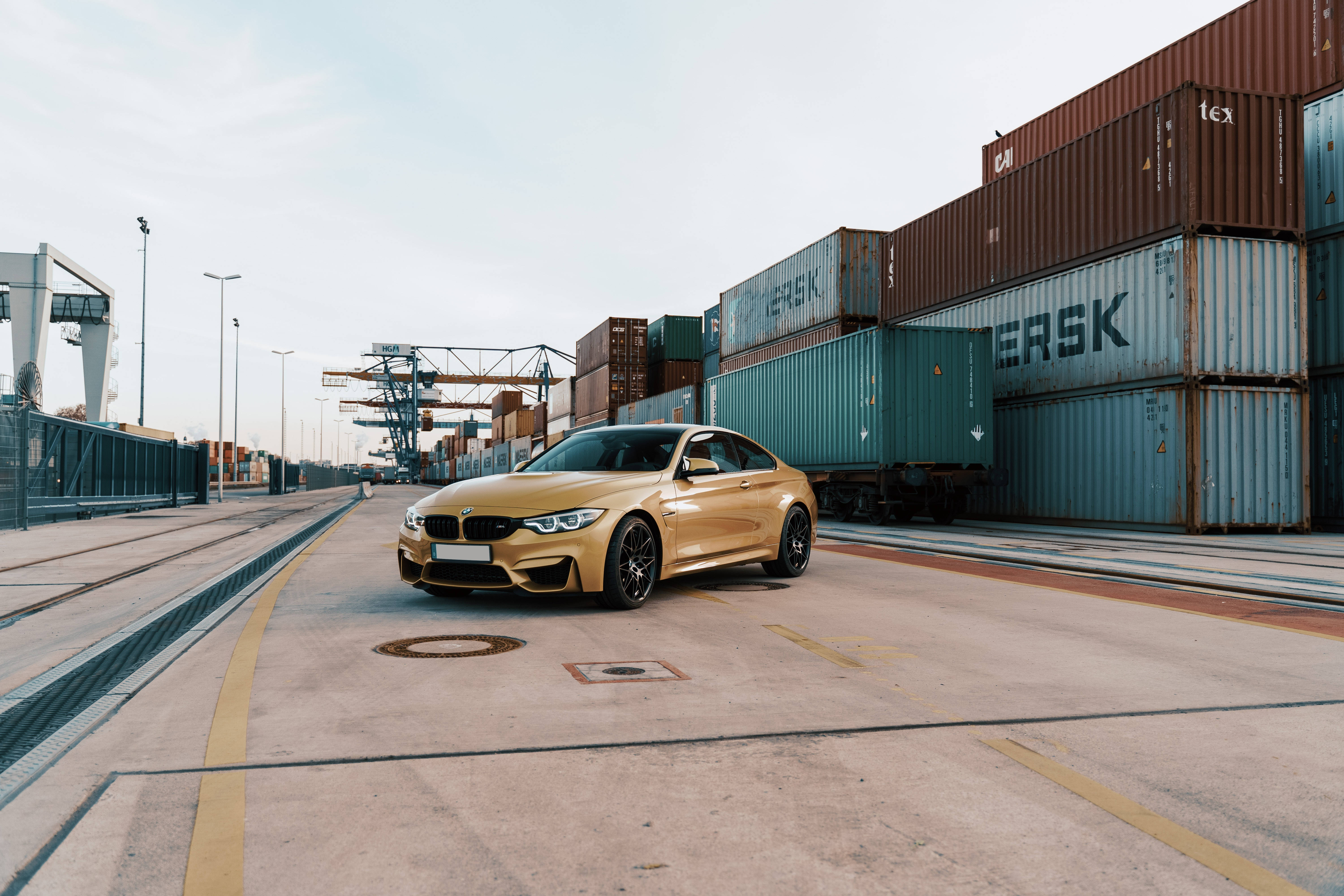 米色 桌布 壁紙 — 一輛閃亮的金色BMW M4雙門跑車停泊在繁忙港口的水泥地上，旁邊是堆疊的貨運集裝箱。