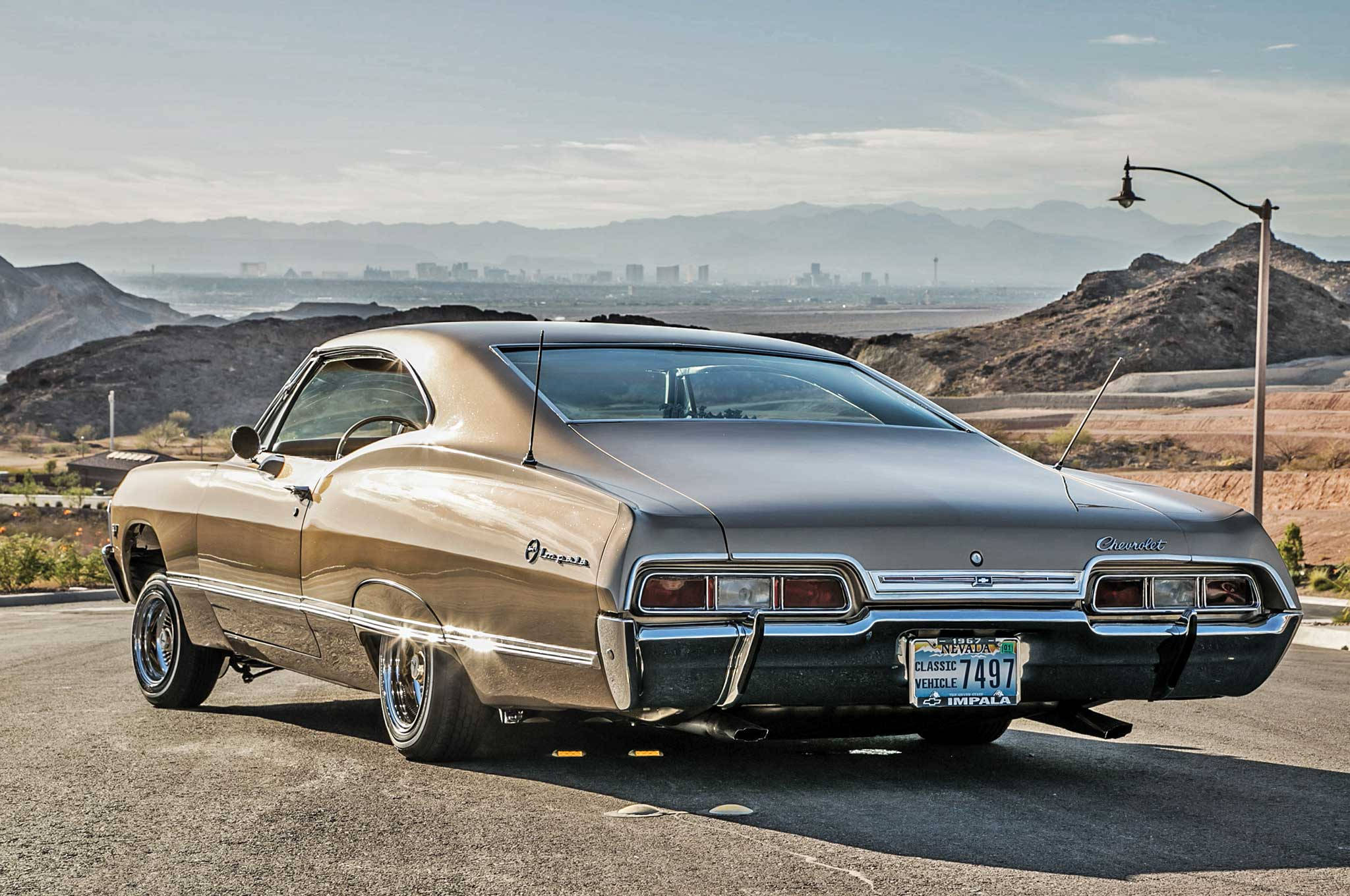 chevrolet impala 1967 桌布 壁紙 — 一輛閃亮的金色經典雪佛蘭 Impala 停泊在沙漠公路上，遠方有山脈和城市天際線，天空朦朧。