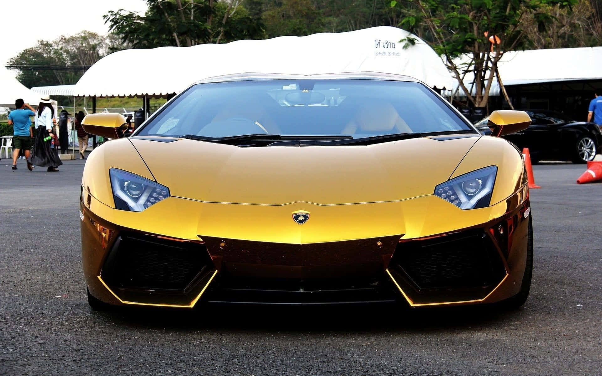 Gold Lamborghini 桌布 壁紙 — 一輛閃亮的金色藍寶堅尼Aventador跑車停放在戶外，背景有人群和白色帳篷。