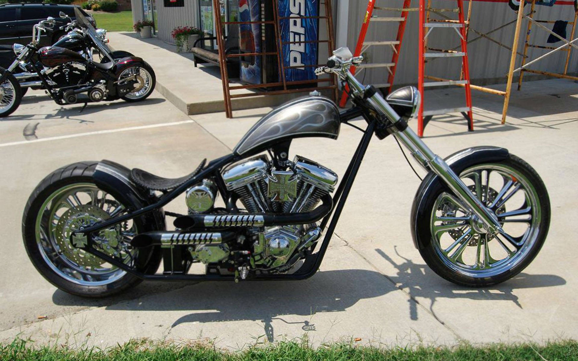 Choppers 桌布 壁紙 — 一輛閃亮的鉻合金訂製chopper摩托車停在混凝土場地上，背景有另一輛摩托車和鷹架。